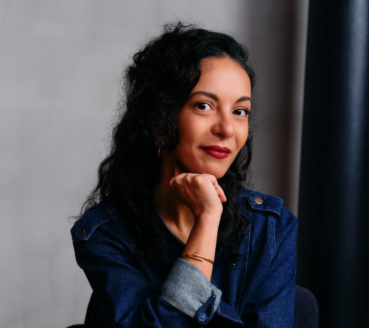 Une femme aux cheveux noirs bouclés, vêtue d'une tenue en jean bleu, est assise sur une chaise, le menton posé sur sa main, et regarde la caméra avec un léger sourire.