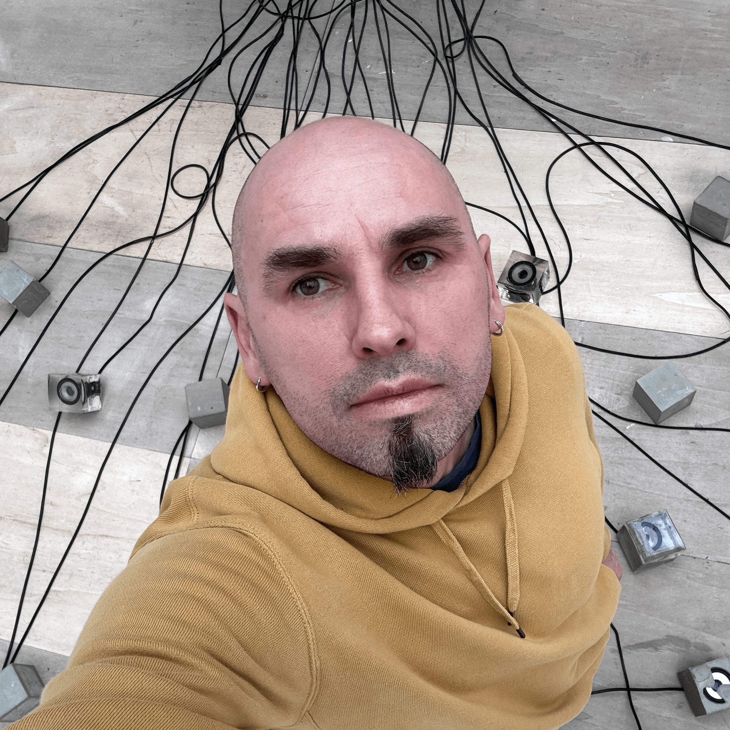 Une personne portant un sweat à capuche jaune se tient sur un sol carrelé, regardant la caméra, entourée de nombreux câbles noirs et de petits appareils gris.