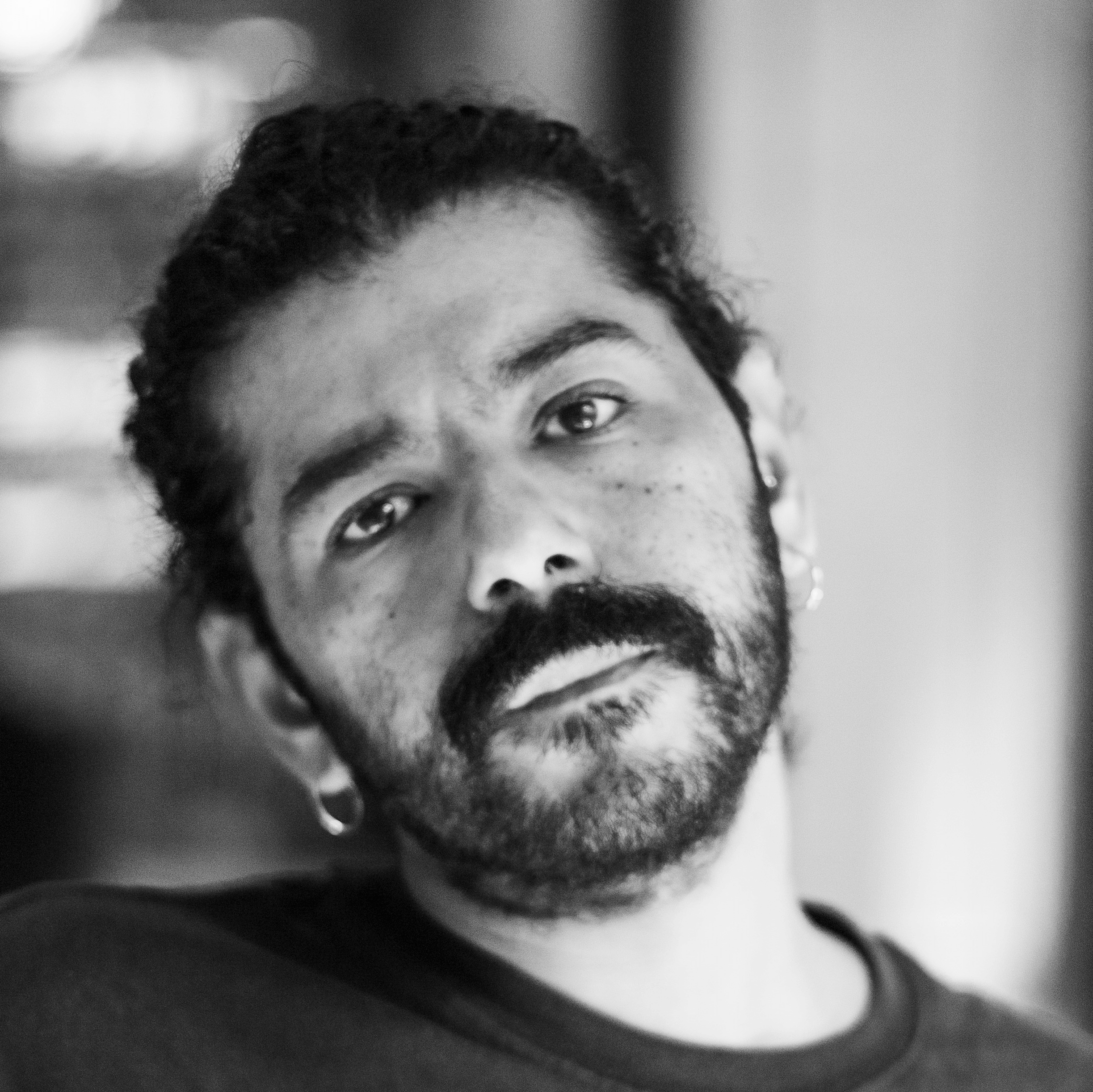 Portrait en noir et blanc d'une personne aux cheveux bouclés, à la barbe et à la moustache, portant des boucles d'oreilles et une chemise sombre, regardant directement l'appareil photo avec une expression neutre.