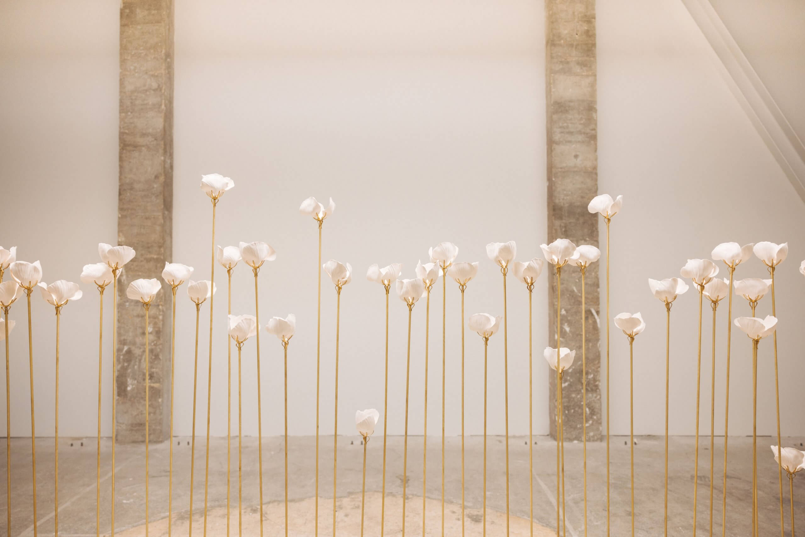 De hautes tiges élancées avec des sculptures blanches ressemblant à des fleurs sont disposées en rangées sur un sol de couleur claire sur un fond minimaliste avec deux colonnes verticales en béton.