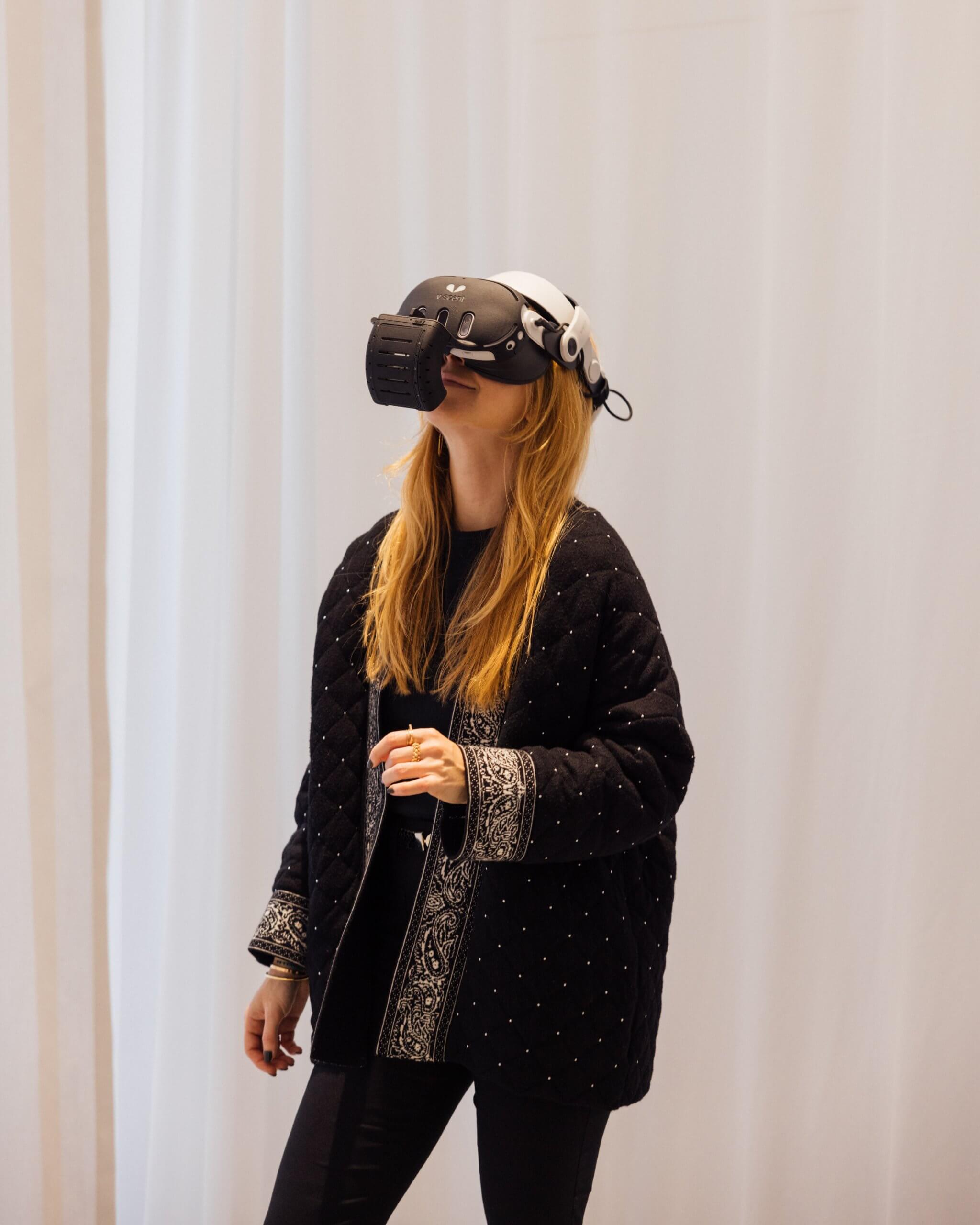 Une femme portant une veste noire à motifs et un casque de réalité virtuelle se tient à l'intérieur devant un rideau blanc.