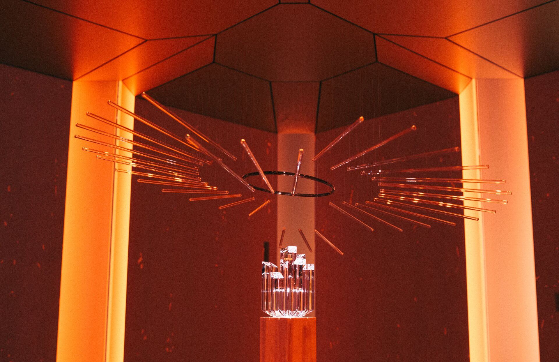 Un luminaire géométrique avec des tiges rayonnantes et un anneau central est suspendu dans une pièce moderne éclairée à l'orange avec des surfaces réfléchissantes.