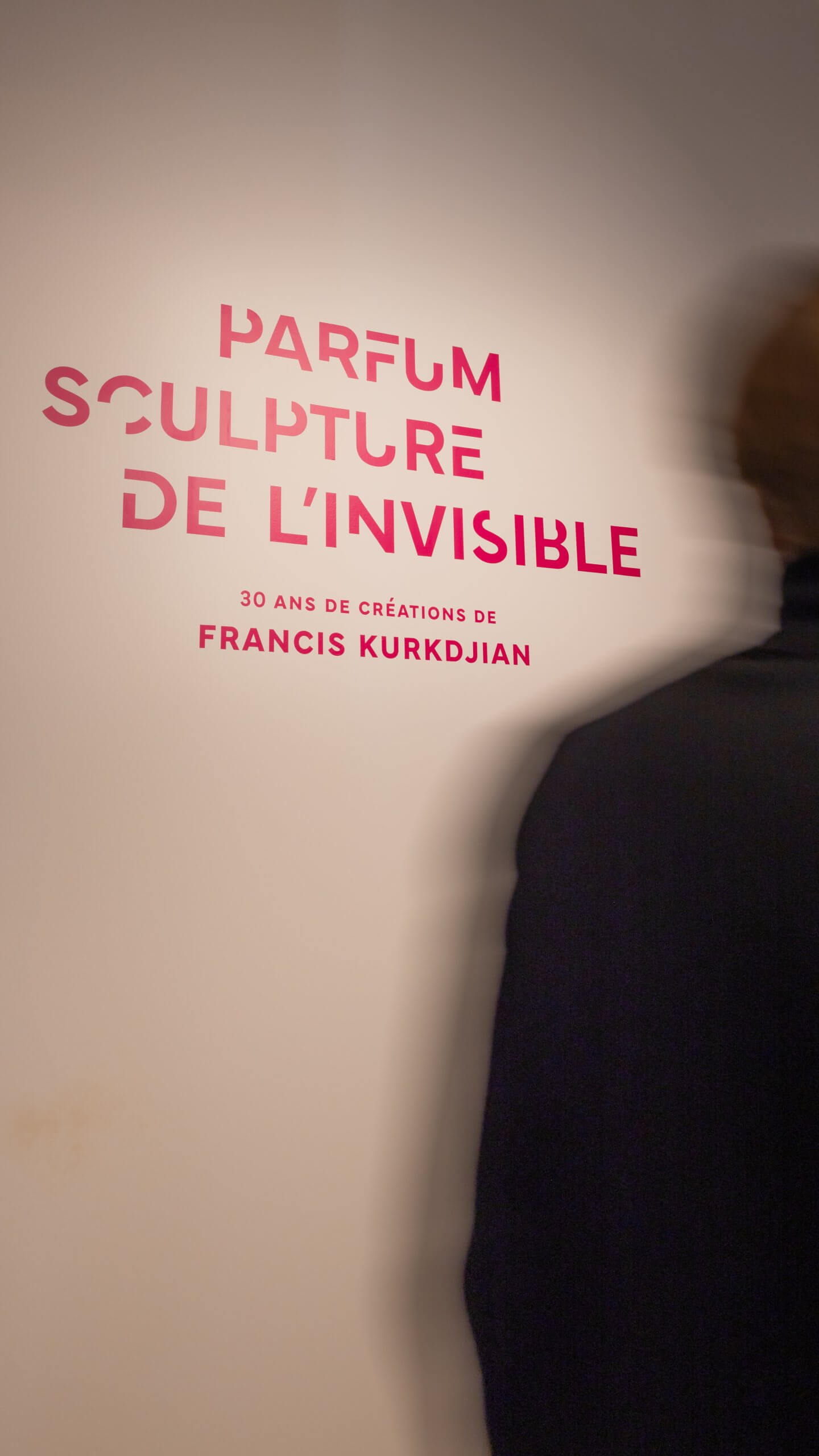 Une silhouette floue se tient près d'un mur sur lequel on peut lire en rose Parfum Sculpture de l'Invisible, 30 ans de créations de Francis Kurkdjian.