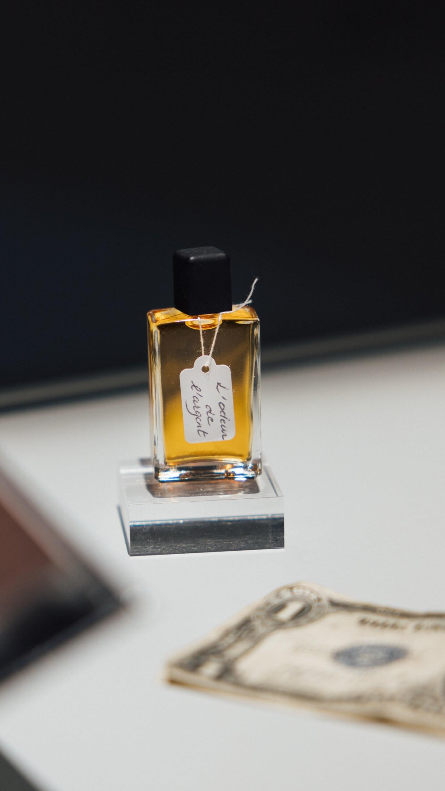 Un flacon de parfum en verre contenant un liquide ambré et une étiquette manuscrite indiquant "L'odeur de l'argent" est posé sur un support près d'un billet de banque américain.