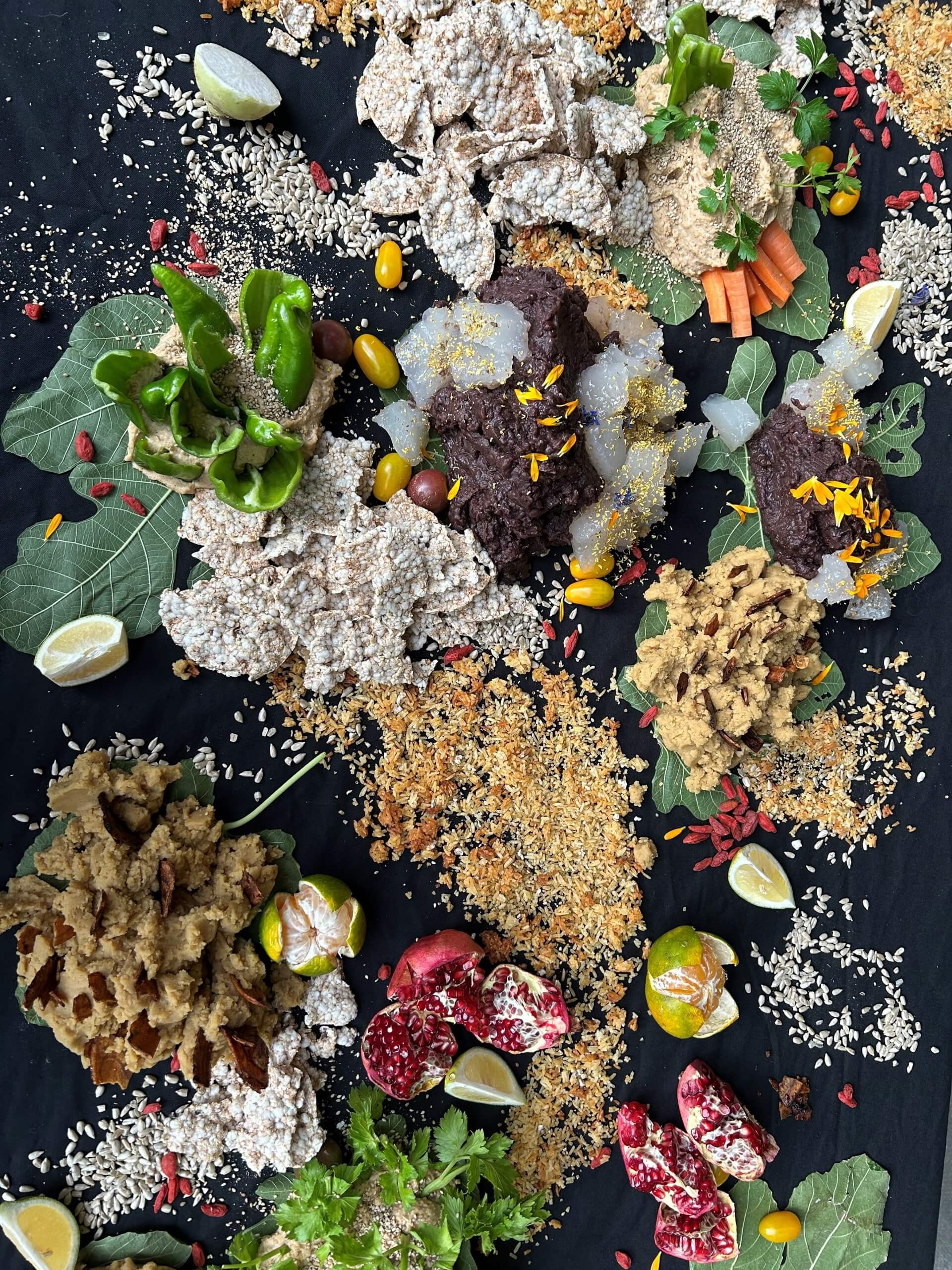 Une variété de plats à base de plantes, de graines, de légumes, de feuilles vertes, de moitiés de grenade et de quartiers de citron disposés sur une surface noire.