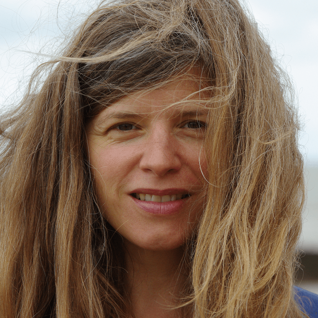 Une personne aux cheveux longs, ondulés et châtains clairs se tient à l'extérieur, regardant l'appareil photo avec une expression détendue.
