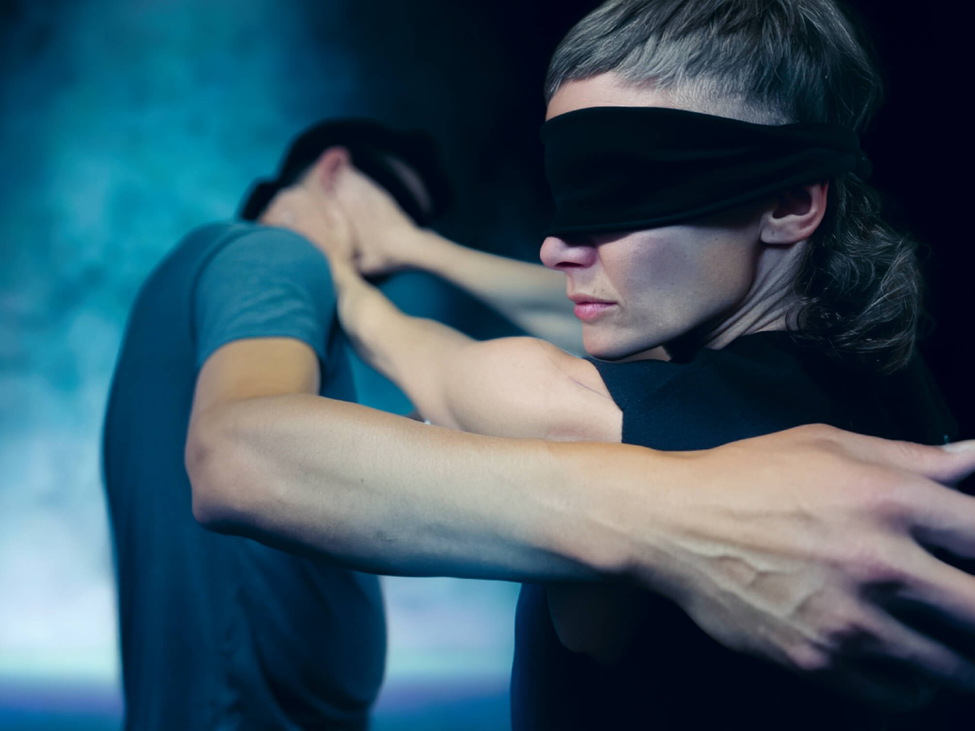 Deux personnes, dont l'une a les yeux bandés de noir, tendent les bras l'une vers l'autre sur un fond flou et coloré.