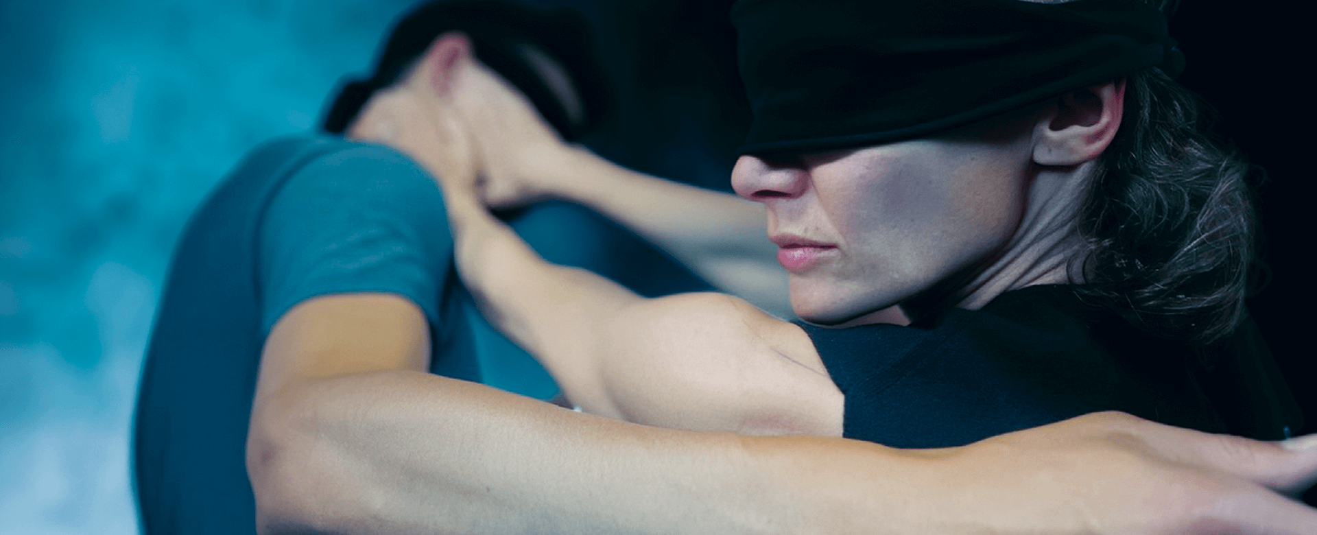 Deux personnes aux yeux bandés, un homme et une femme, se tiennent l'un près de l'autre, les bras tendus, semblant se livrer à une activité physique ou à une danse.