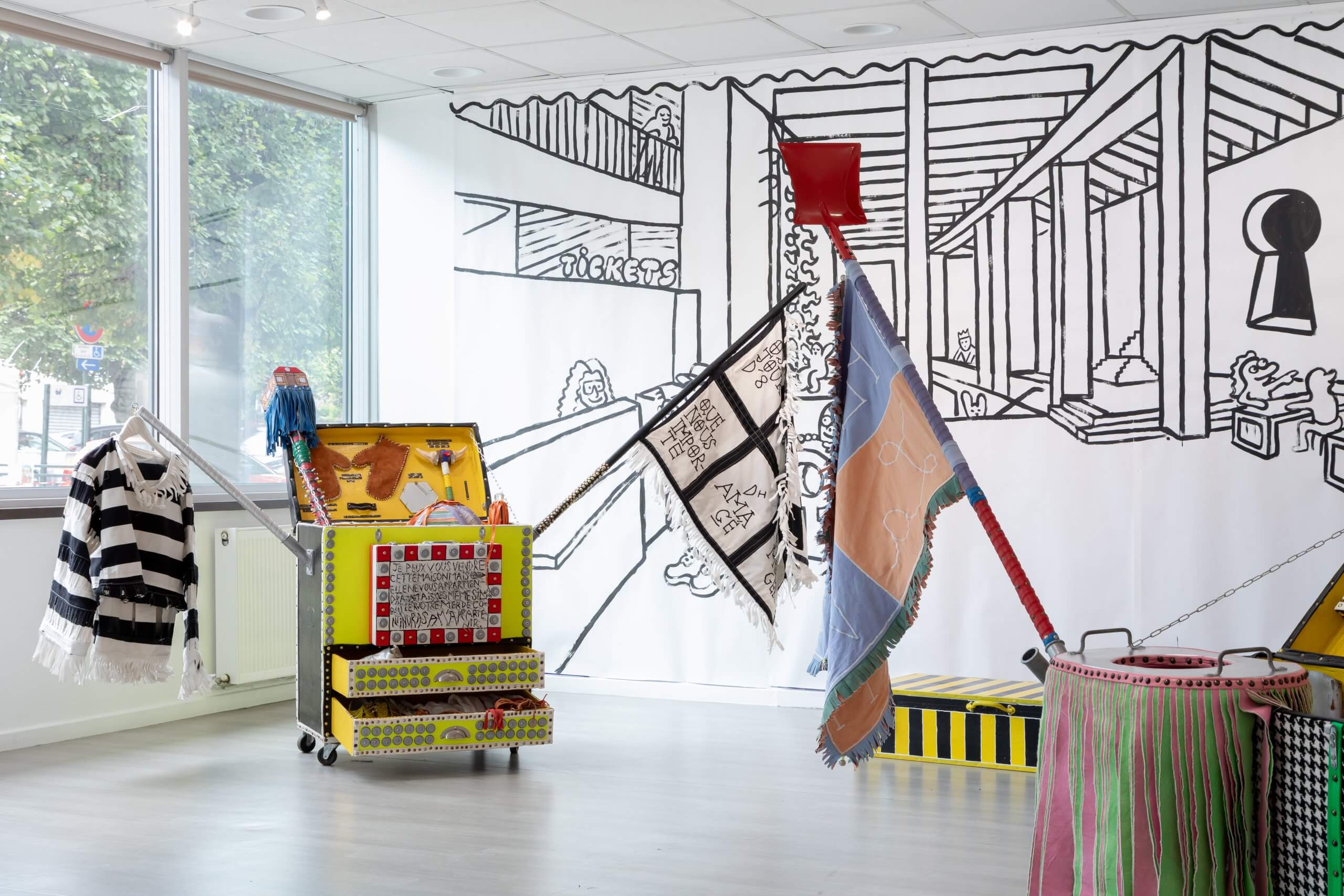 Une salle très éclairée avec des chariots décorés, des drapeaux colorés et des costumes devant une grande peinture murale en noir et blanc représentant une scène architecturale.