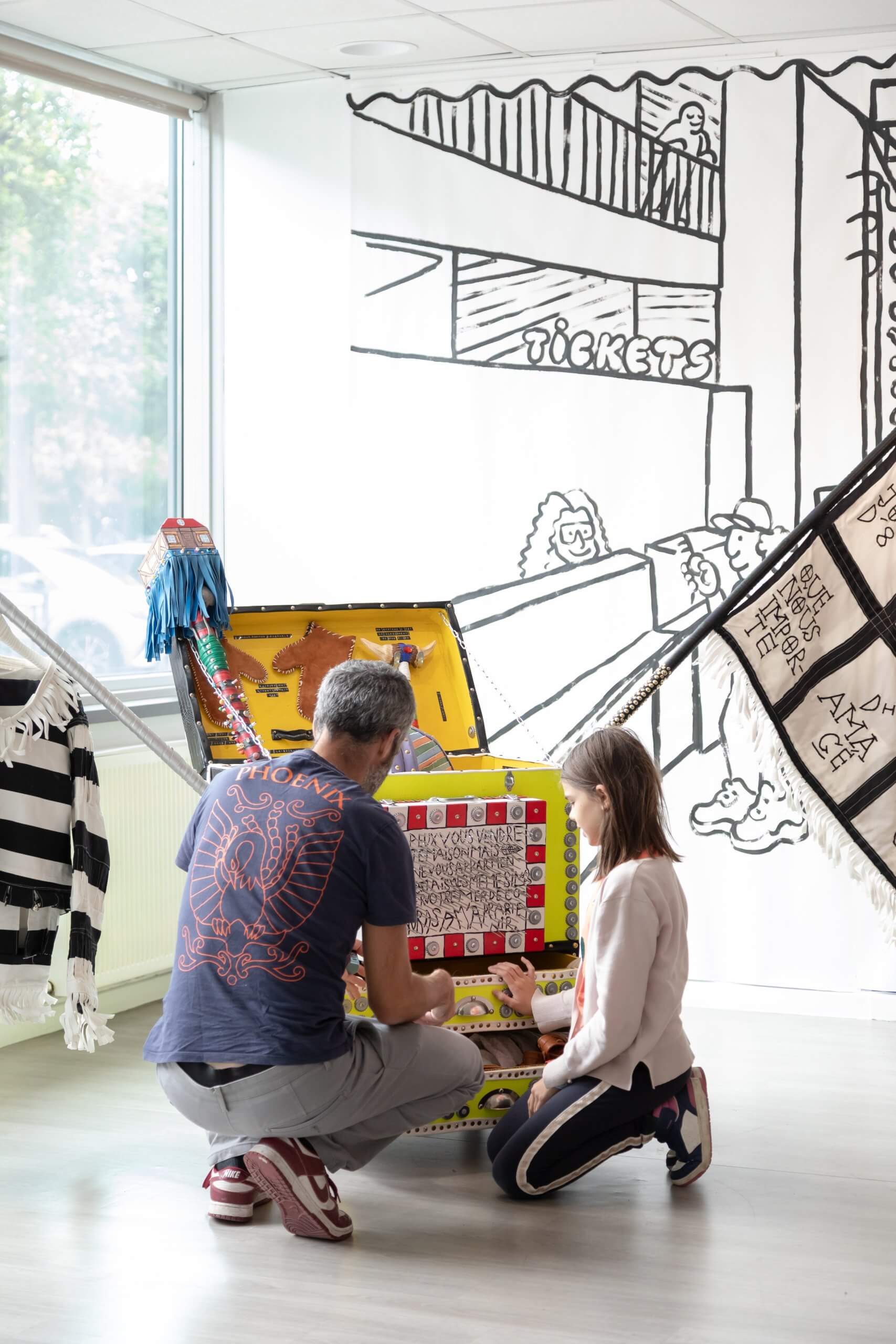 Un adulte et un enfant sont agenouillés devant une boîte colorée et décorée dans une pièce lumineuse où sont exposés des peintures murales et des costumes.