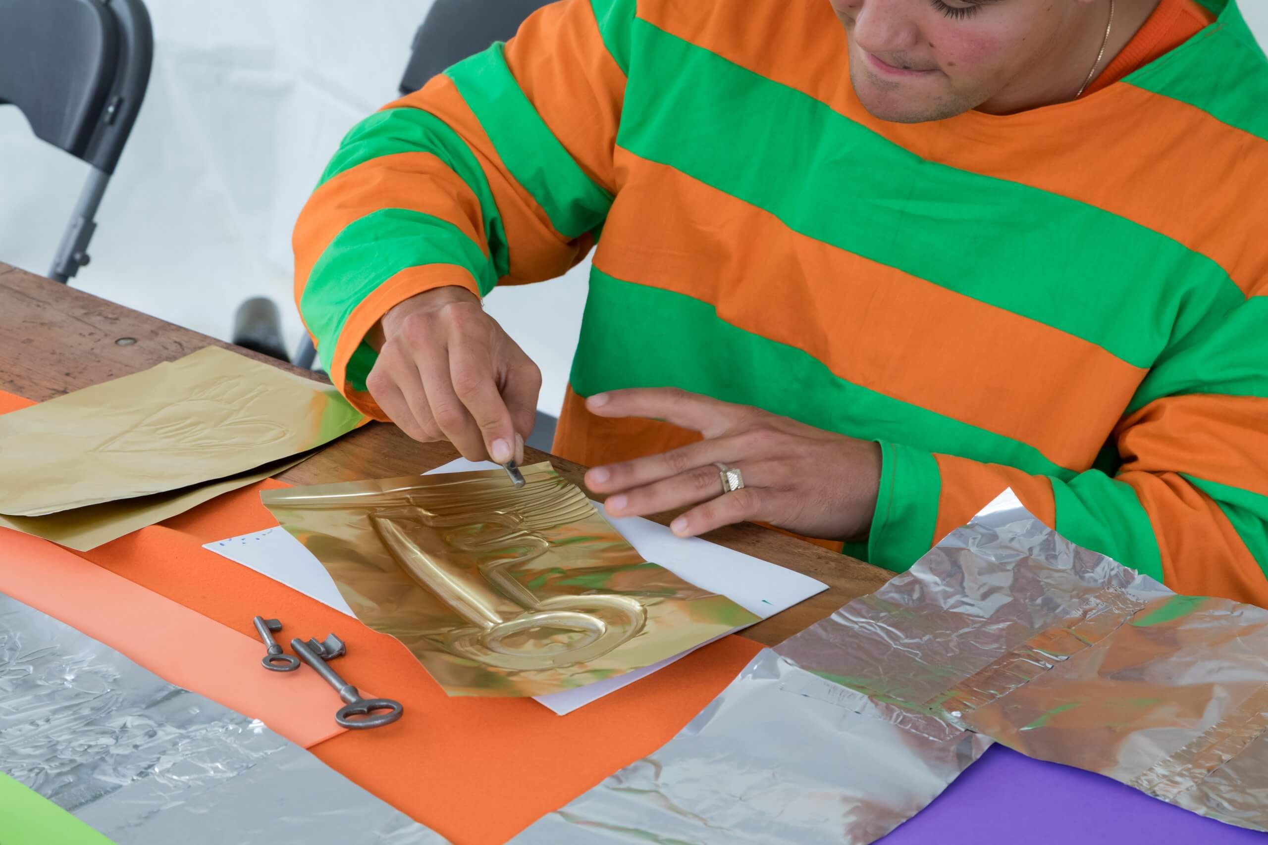 Une personne vêtue d'une chemise à rayures vertes et orange gaufre une forme de clé sur une feuille d'or à l'aide d'un stylet, à une table où se trouvent des papiers colorés et des clés en métal.