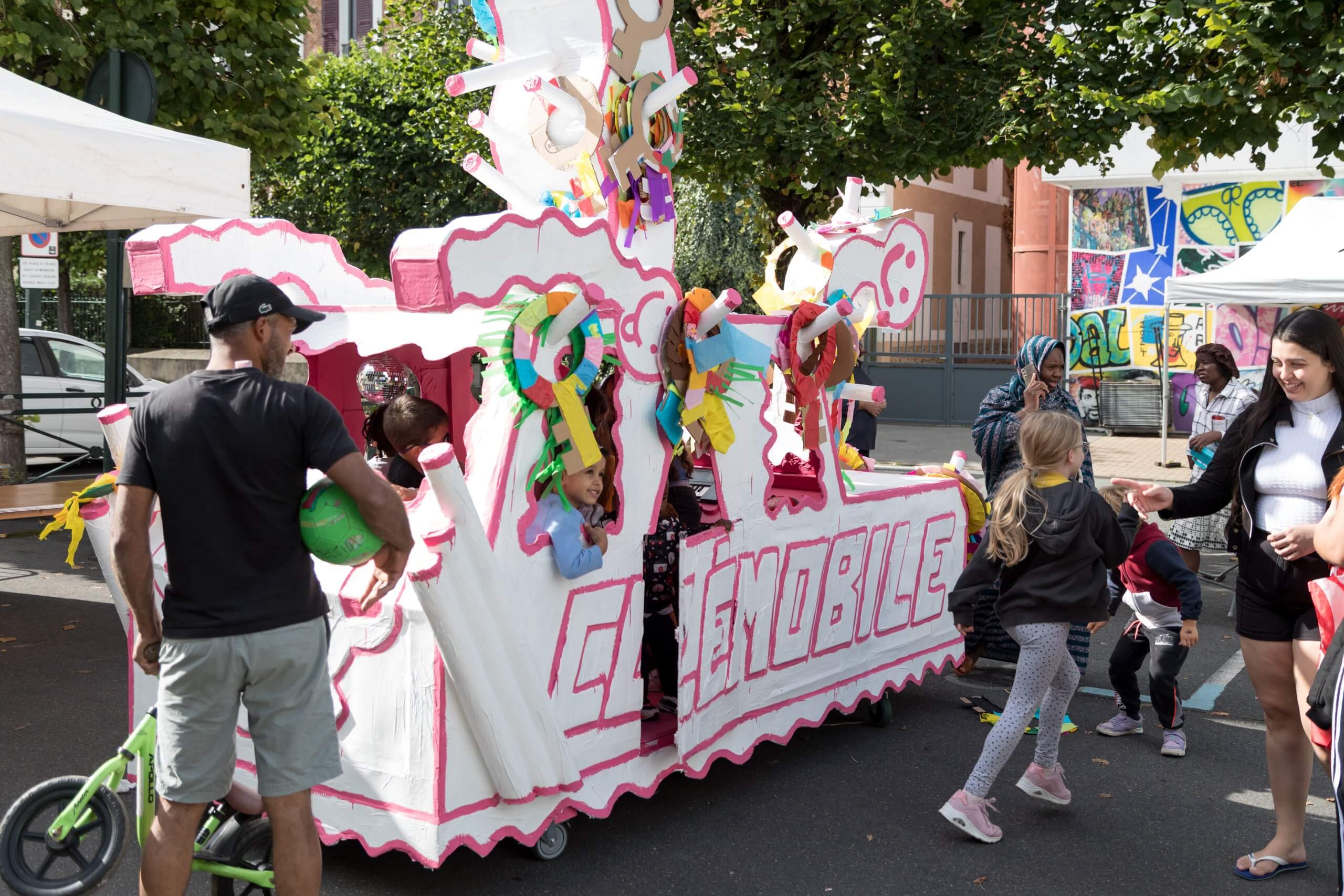 Un groupe de personnes et d'enfants se rassemblent autour d'un véhicule en carton coloré et décoré, étiqueté CANEMOBILE, lors d'un événement en plein air.