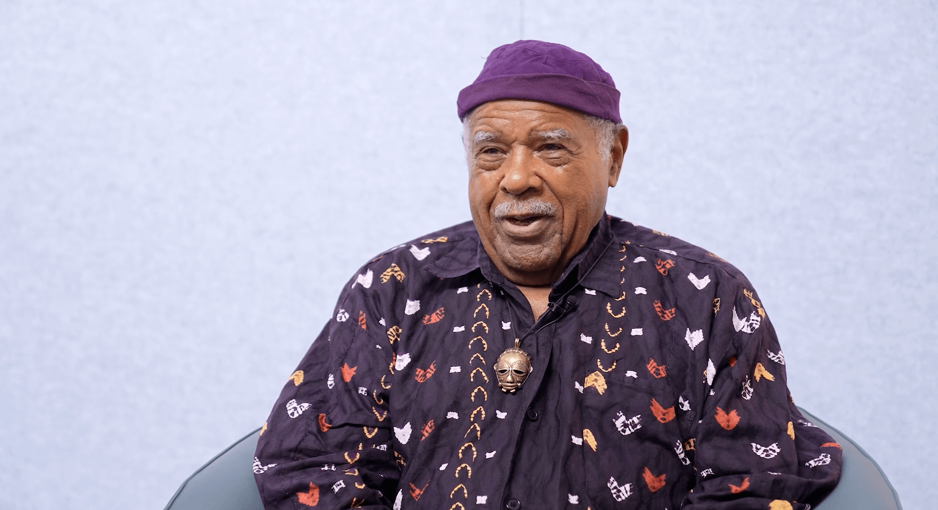 An older man sits in a chair, wearing a purple patterned shirt and matching cap, against a plain light gray background.