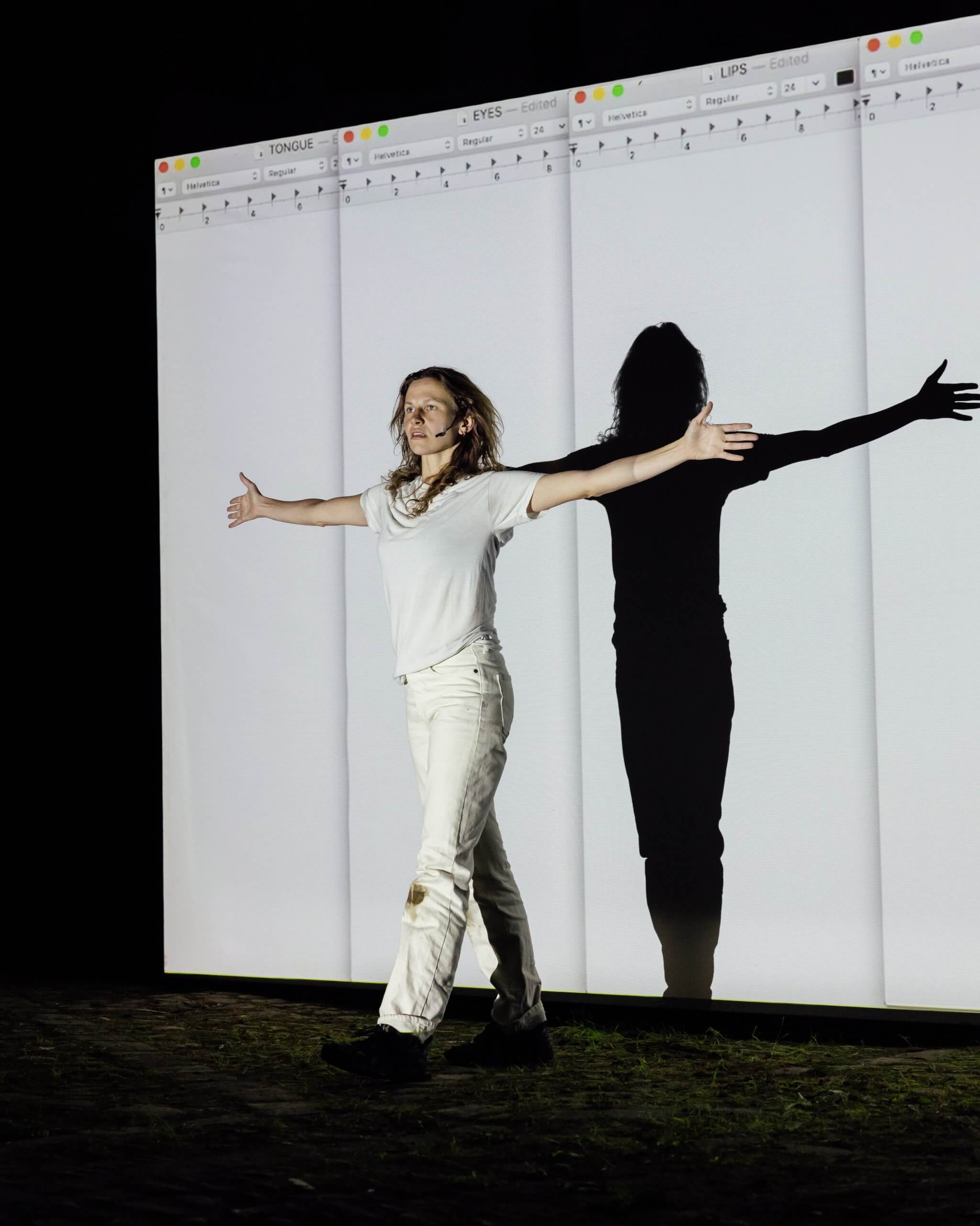 Une personne vêtue de blanc se tient debout, les bras tendus, devant un grand écran affichant des feuilles de calcul, projetant une ombre.