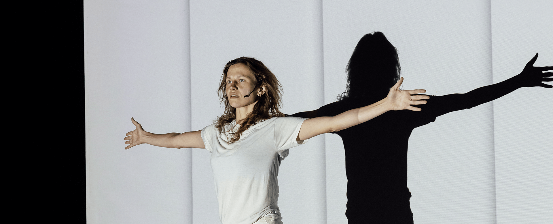 Une personne vêtue d'une chemise blanche se tient debout, les bras tendus, devant une toile de fond blanche, projetant une ombre ; elle porte un micro-casque.