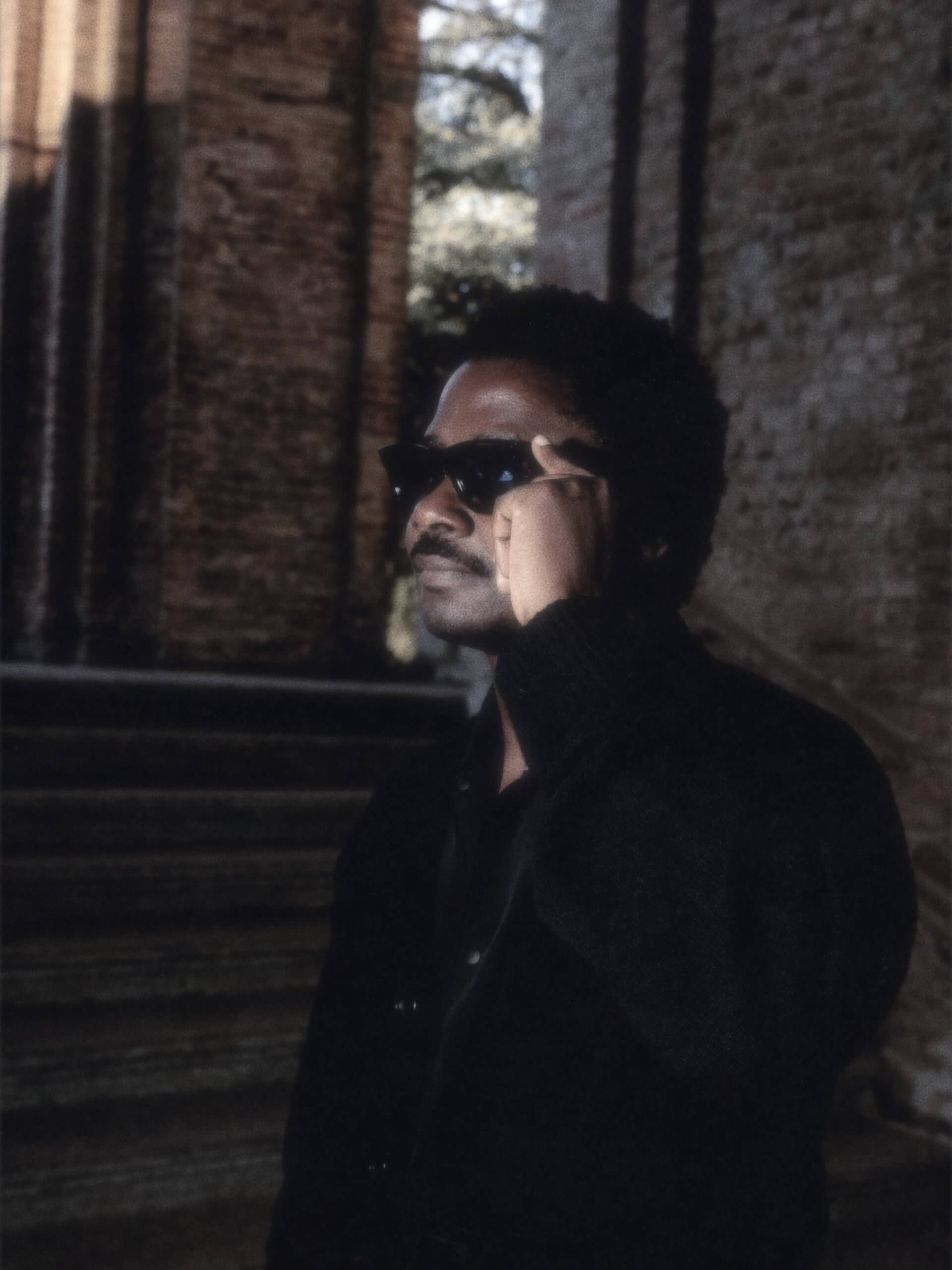 A person wearing dark sunglasses and a dark jacket stands in front of stone steps and a brick wall, holding their glasses with one hand.