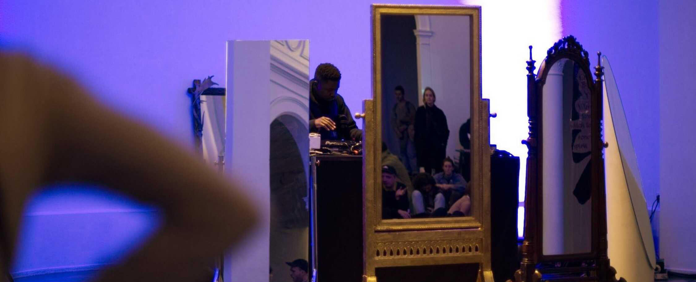 Plusieurs personnes se reflètent dans des miroirs ornés placés dans une pièce faiblement éclairée par des lumières bleues et violettes ; une personne se tient à côté des miroirs.