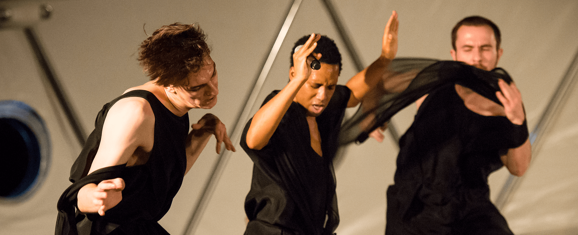 Trois personnes vêtues de noir exécutent une danse contemporaine expressive, chacune tenant un tissu transparent et se déplaçant énergiquement sur un fond clair.