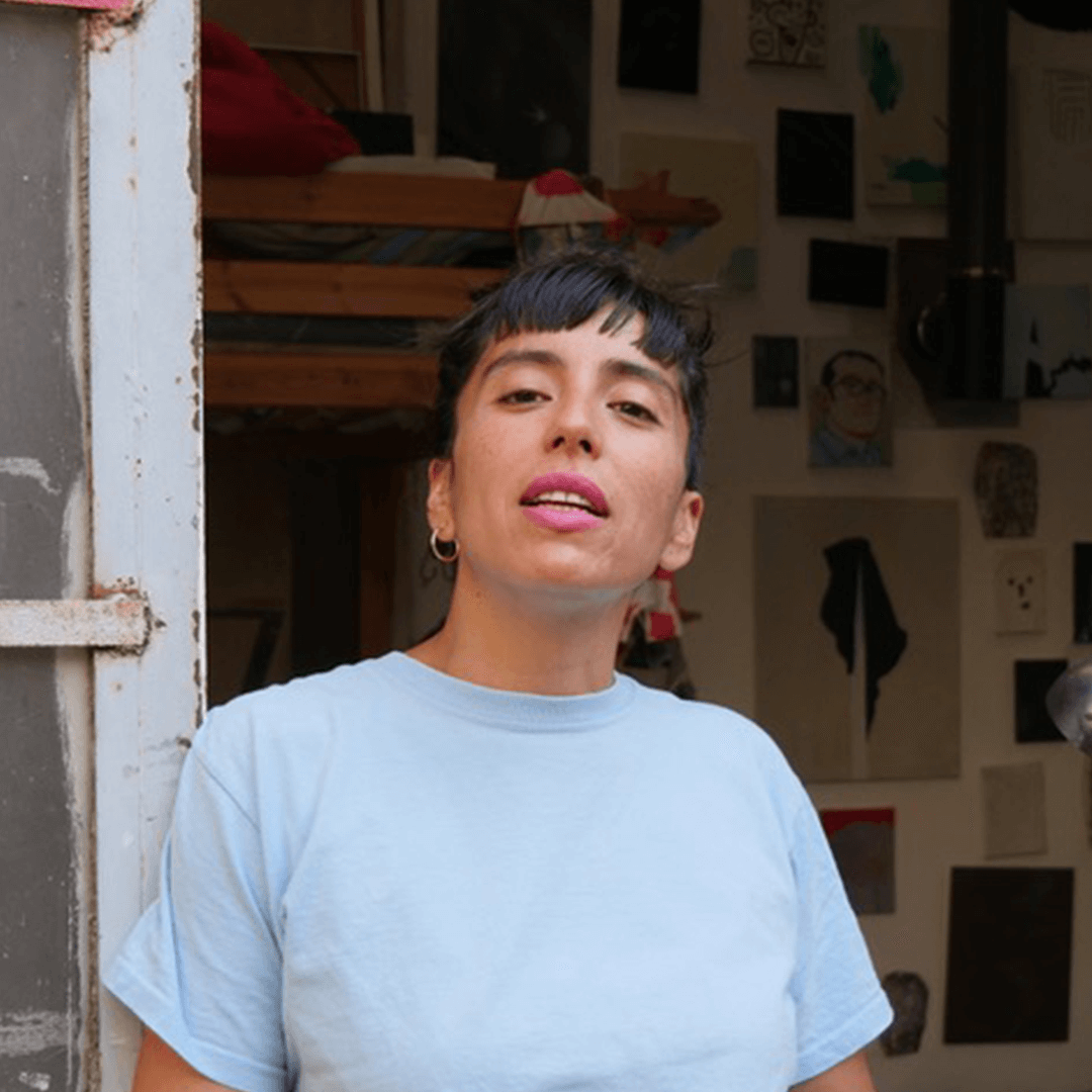 Une personne aux cheveux bruns courts et à la chemise bleu clair se tient devant une porte ouverte, avec des œuvres d'art affichées sur le mur derrière elle.