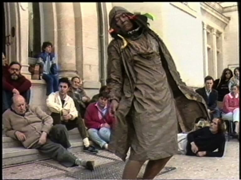Une personne vêtue d'un costume en lambeaux auquel sont attachés des morceaux de tissu se tient à l'extérieur, près d'un bâtiment en pierre clair ; une autre personne regarde par une fenêtre voisine.