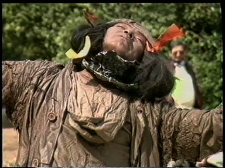 Une personne aux cheveux longs, aux vêtements tachés de boue et aux rubans colorés dans les cheveux se tient à l'extérieur, la tête penchée en arrière et les bras tendus. Une autre personne est visible à l'arrière-plan.