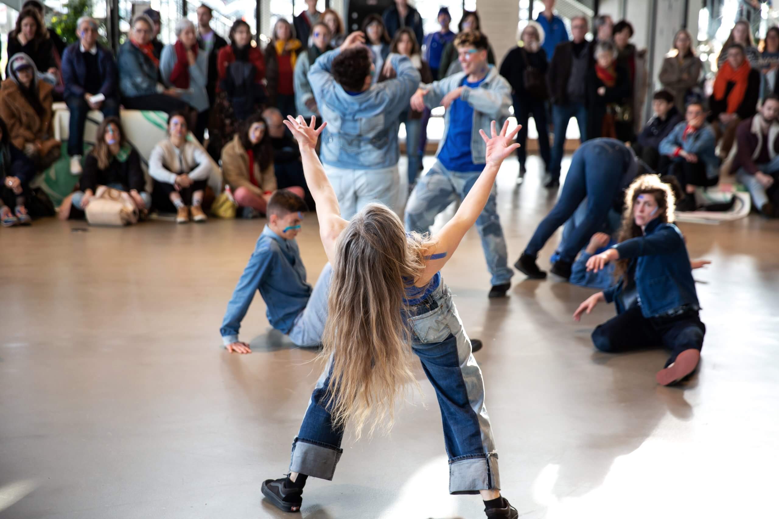 Un groupe de danseurs vêtus de tenues en jean s'exécute énergiquement sur un sol ciré, tandis qu'un public assis regarde en arrière-plan.