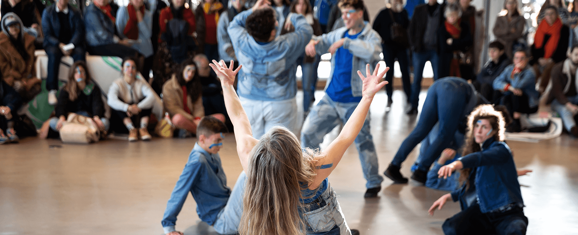 Un groupe de personnes portant des jeans exécute une danse au centre d'une grande salle, tandis qu'un public assis et debout autour d'eux les observe.