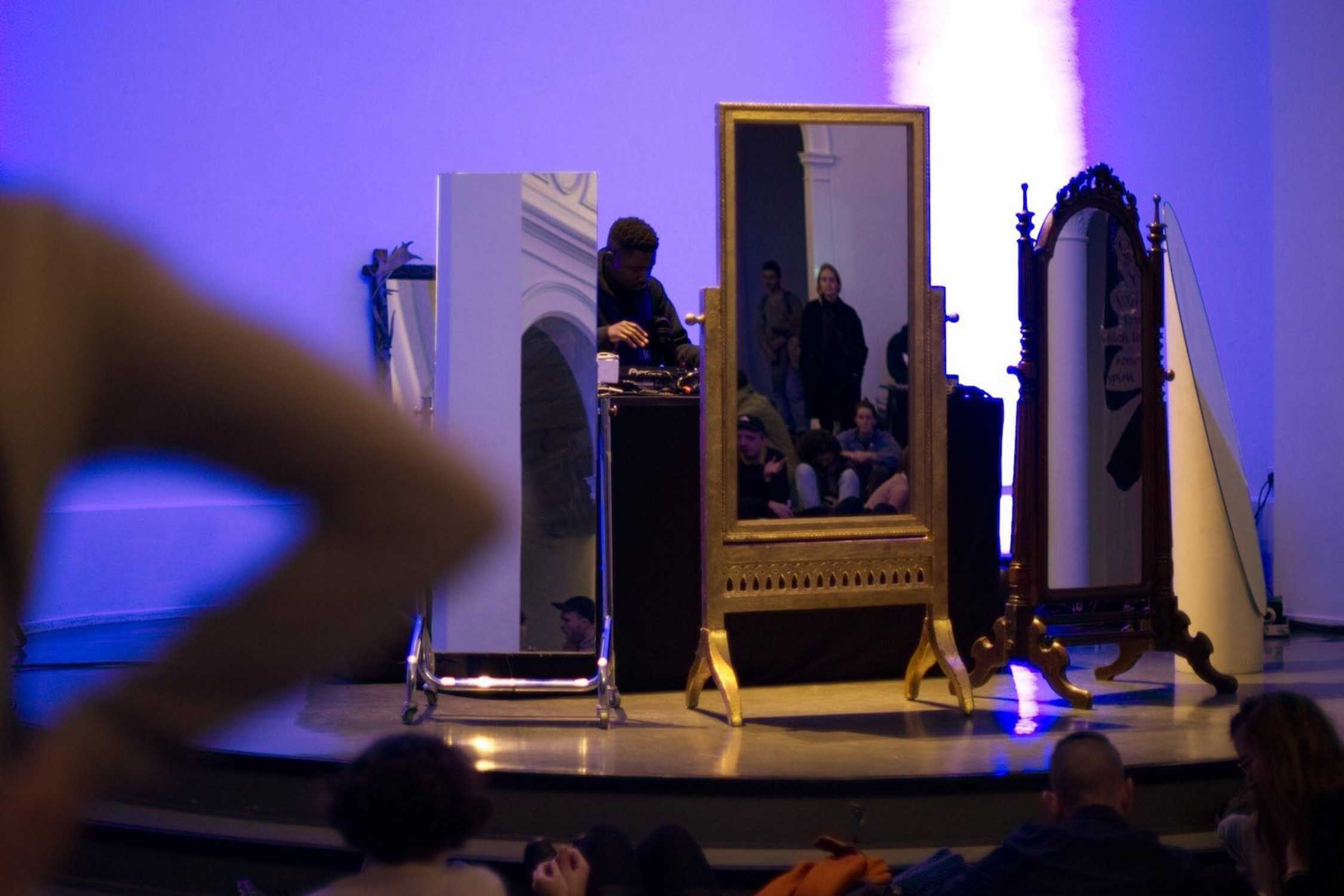 A person stands at a table on a stage surrounded by three large mirrors, with people reflected and seated nearby under blue lighting.