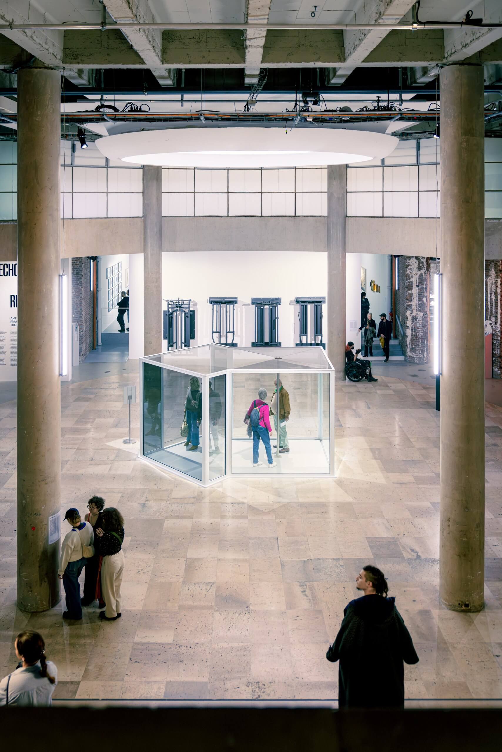 Les visiteurs observent et se promènent autour d'une installation en forme de cube de verre dans un hall d'exposition spacieux et moderne avec de hauts plafonds et de grandes colonnes.