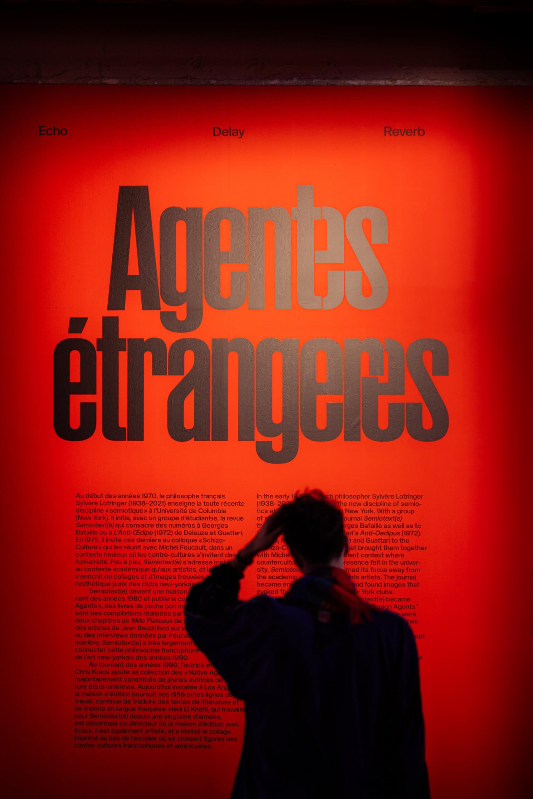 Une personne se tient devant un mur rouge vif sur lequel on peut lire en gros caractères noirs Agents étrangers et, en dessous, des sections de texte plus petites.