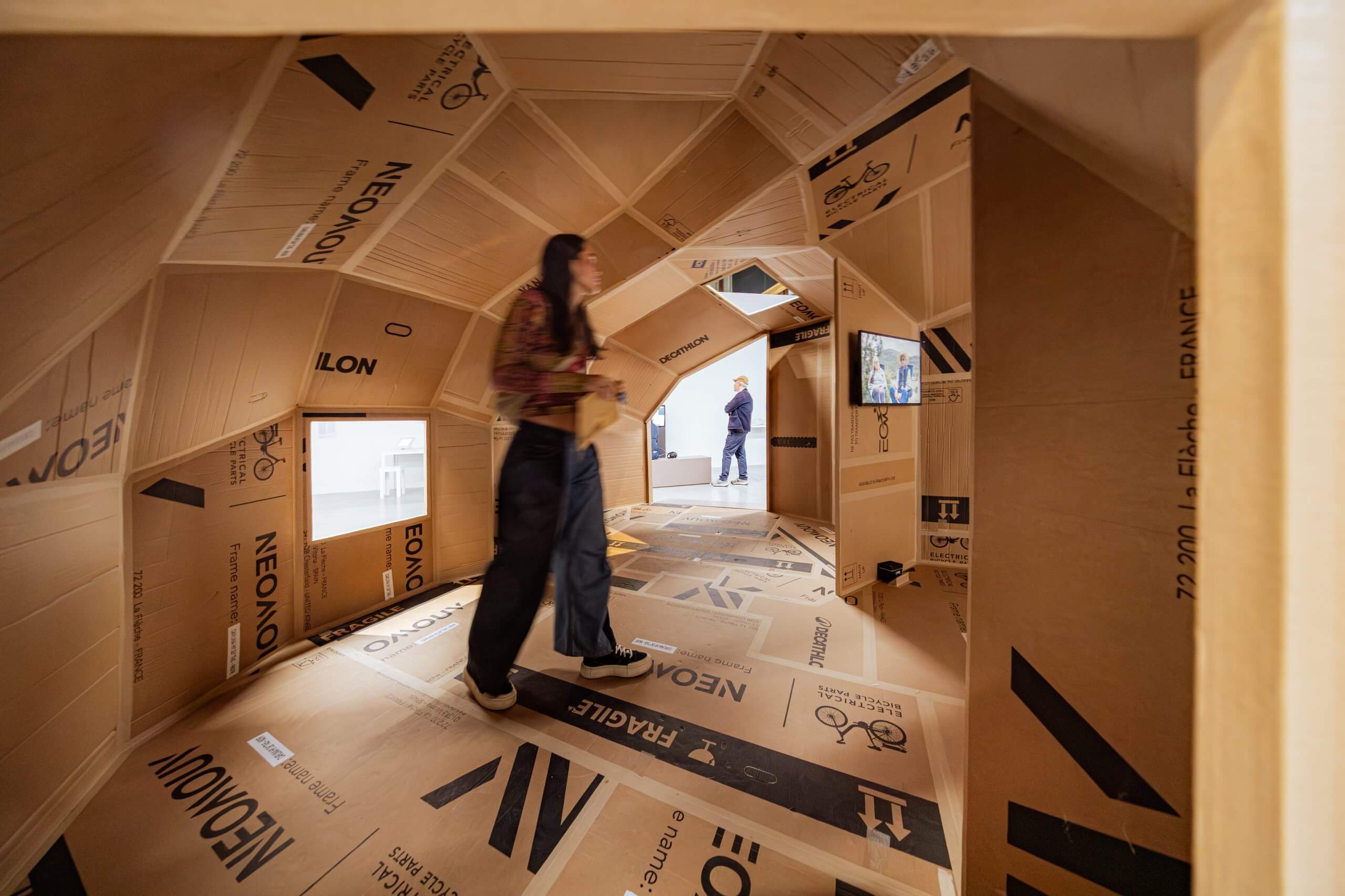 Une personne se tient à l'intérieur d'une structure faite de panneaux de carton avec des textes et des symboles imprimés ; une autre personne est visible à l'extérieur par une ouverture.