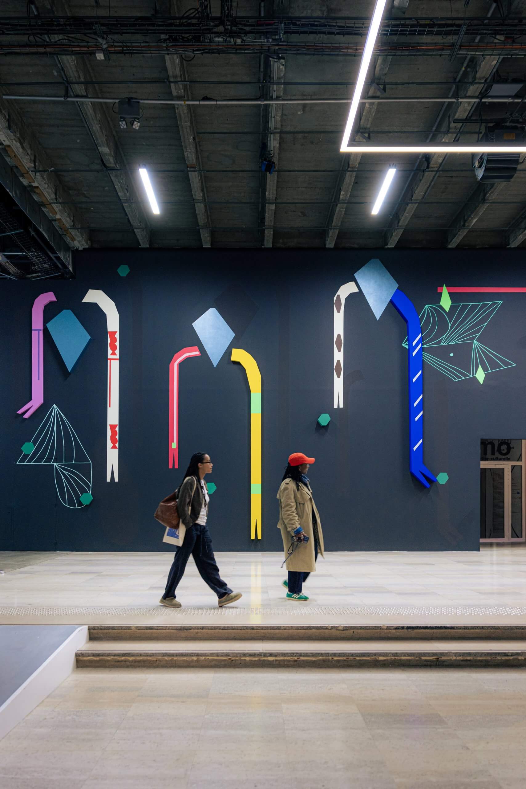 Deux personnes pénètrent dans un espace moderne dont le mur sombre est décoré de grands motifs colorés et abstraits en forme d'alphabets.