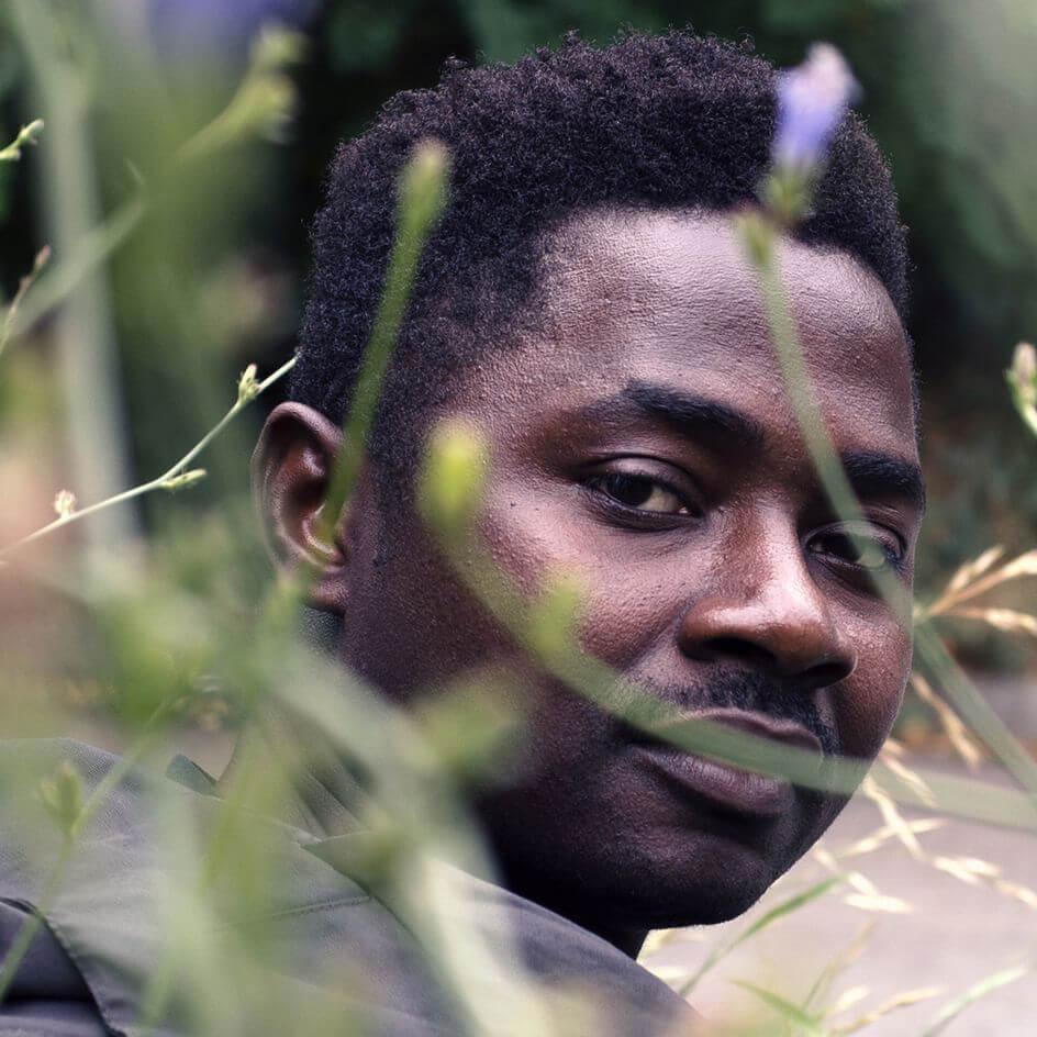 Un homme aux cheveux courts et à la moustache regarde par-dessus son épaule, partiellement caché par des plantes vertes au premier plan.
