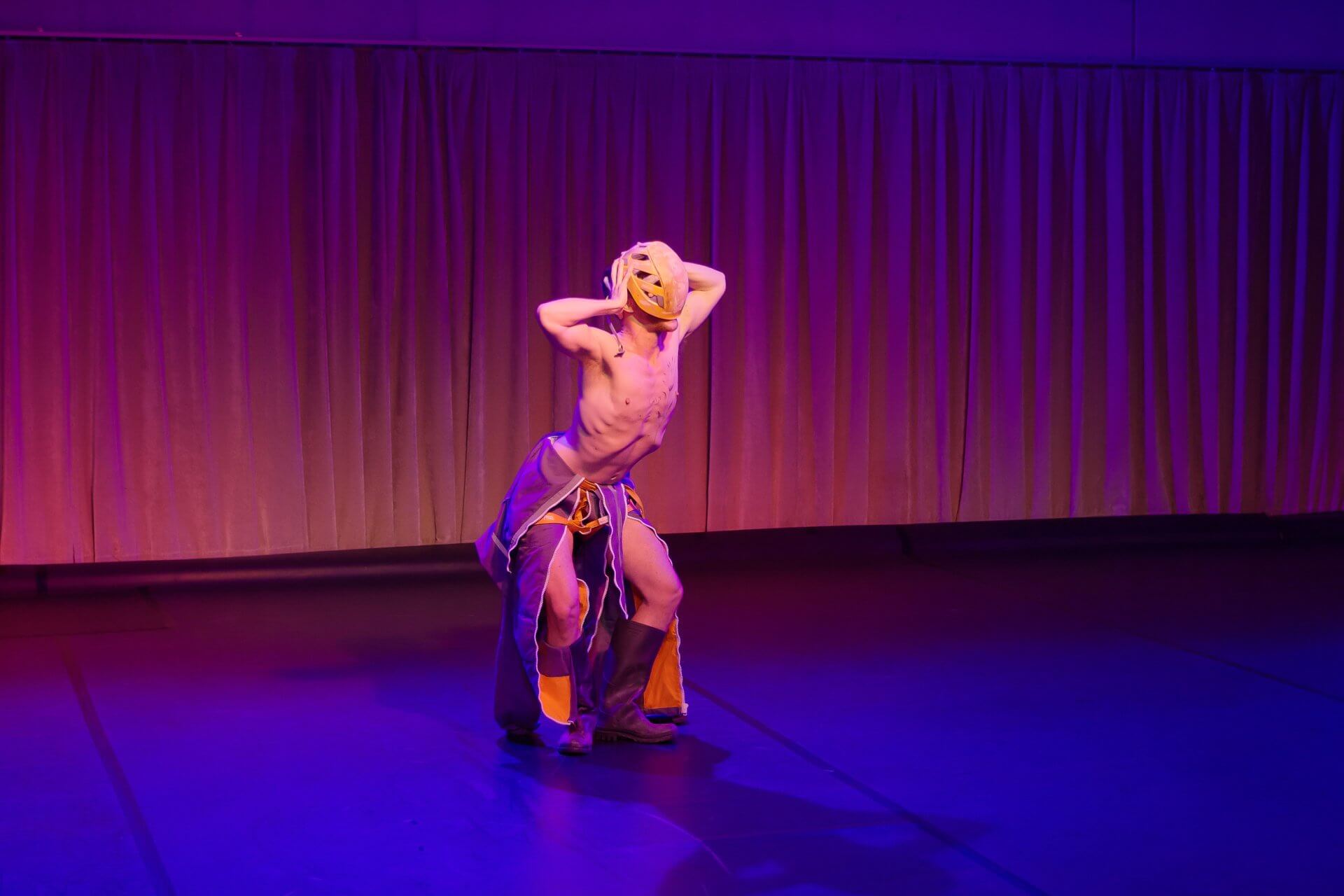 Un artiste sur scène porte un masque et un pantalon ample, et se tient debout dans une pose dramatique, les mains sur la tête, sous un éclairage violet et jaune.