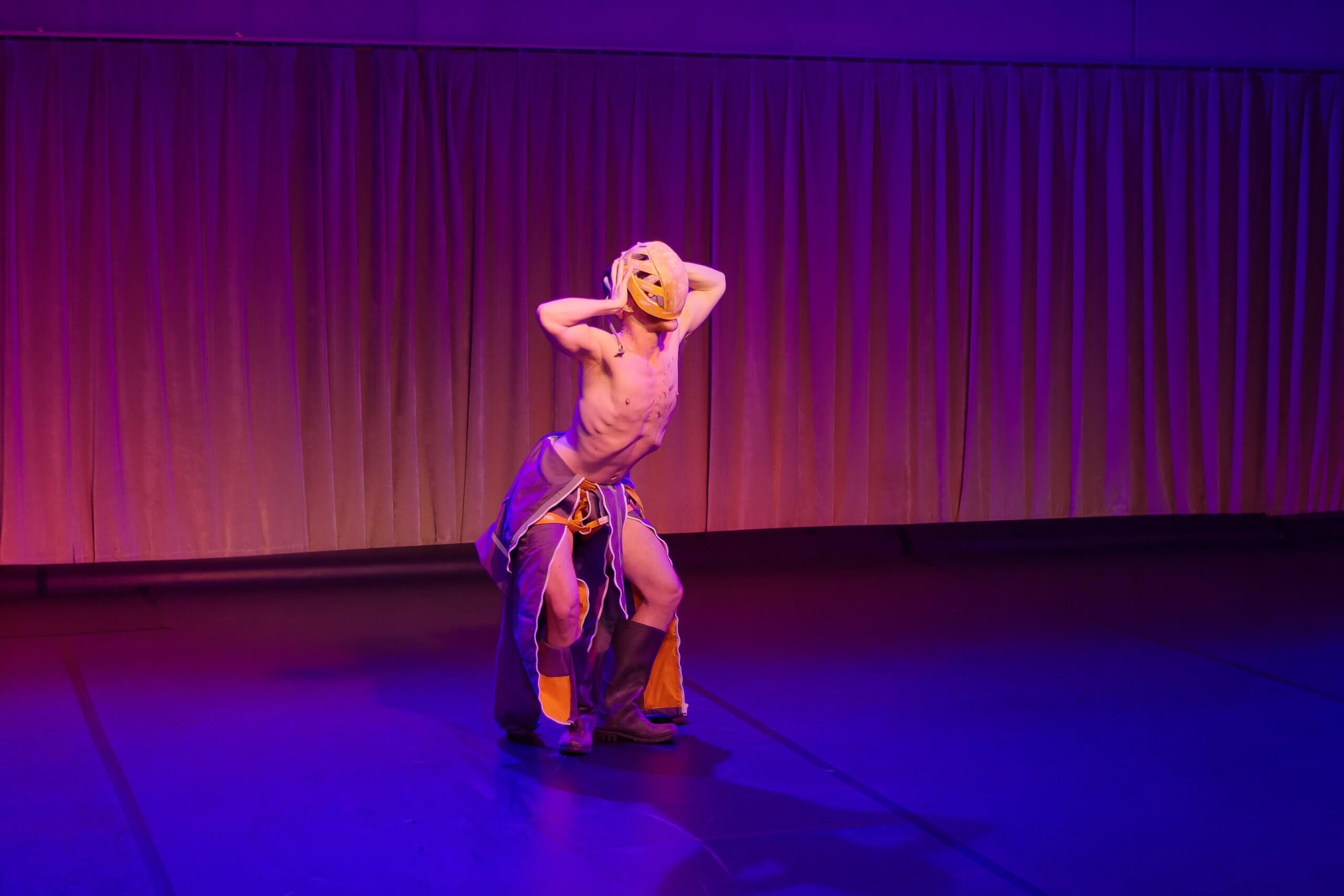 Un artiste sur scène porte un masque et un pantalon ample, et se tient debout dans une pose dramatique, les mains sur la tête, sous un éclairage violet et jaune.