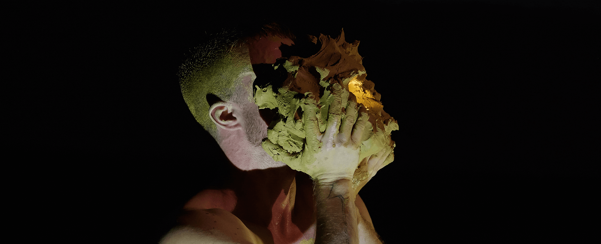 Une personne presse un gros bloc d'argile humide contre son visage et ses mains, avec un éclairage dramatique sur fond noir.