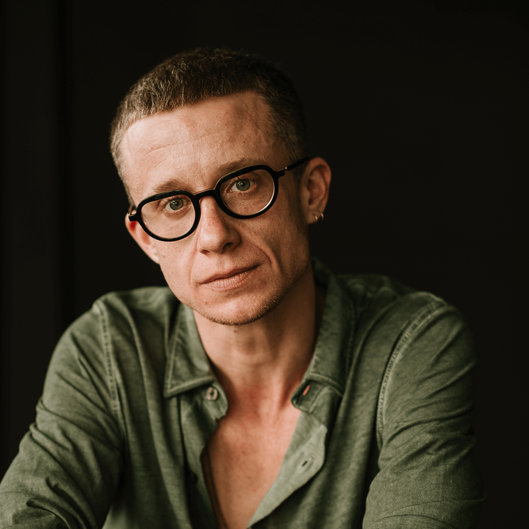 Une personne aux cheveux courts, portant des lunettes et une boucle d'oreille, vêtue d'une chemise verte, est assise sur un fond sombre et regarde directement la caméra.