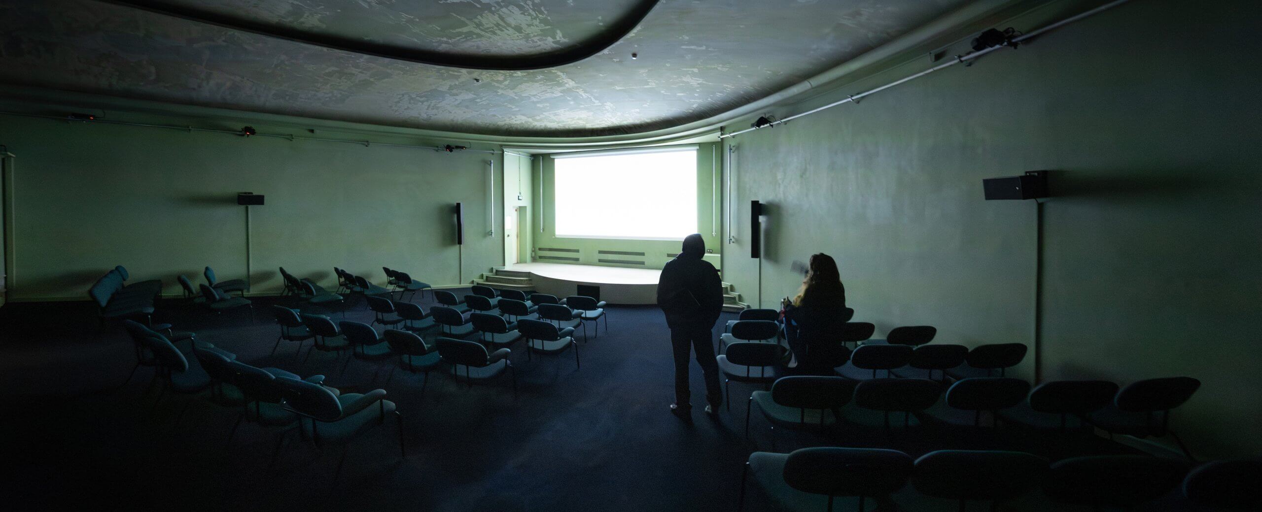 Deux personnes se trouvent dans un théâtre vide et faiblement éclairé, face à un écran blanc et lumineux, entouré de rangées de chaises inoccupées.