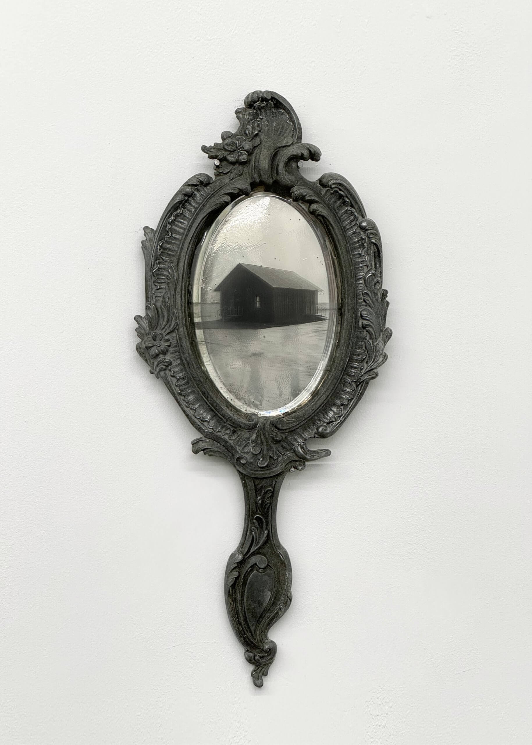 Miroir à main antique orné d'une image réfléchie en noir et blanc d'une petite maison en bois sur un paysage plat.