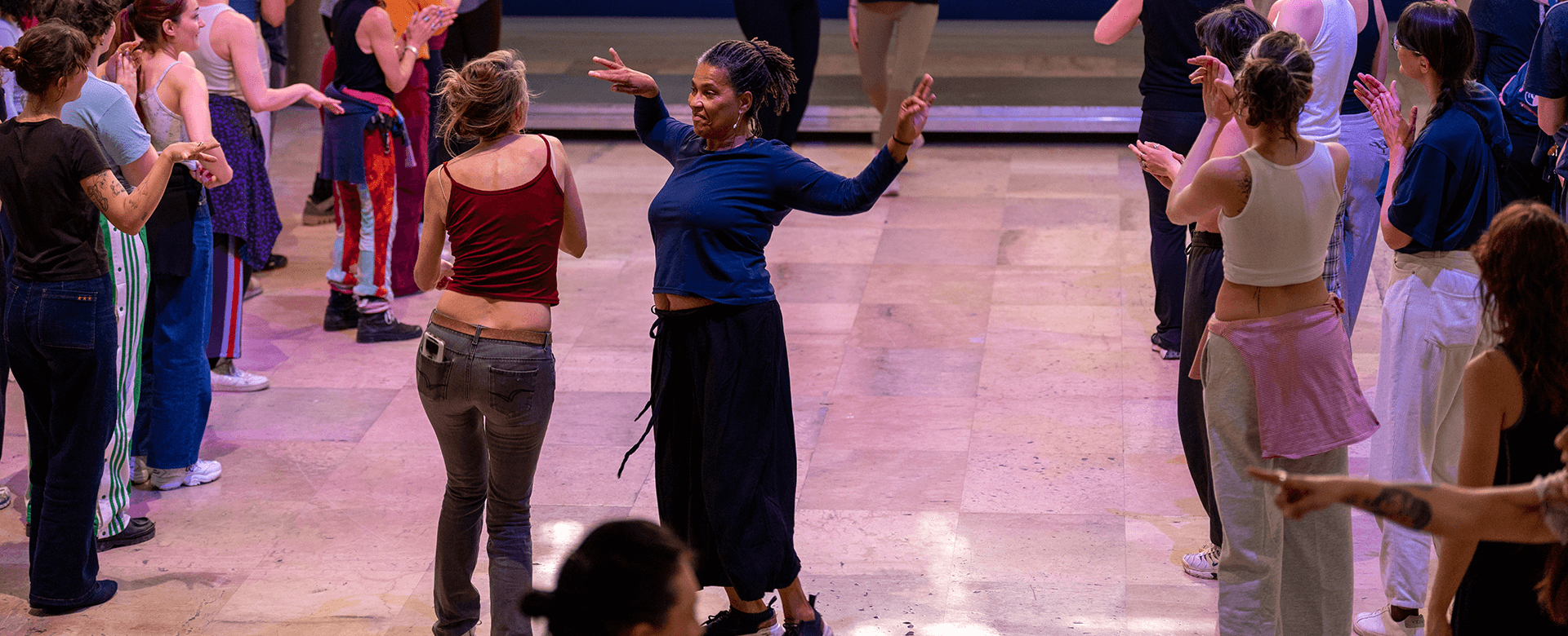 Un groupe de personnes danse ensemble sur un sol ciré à l'intérieur, avec au centre deux femmes qui se font face et se sourient.