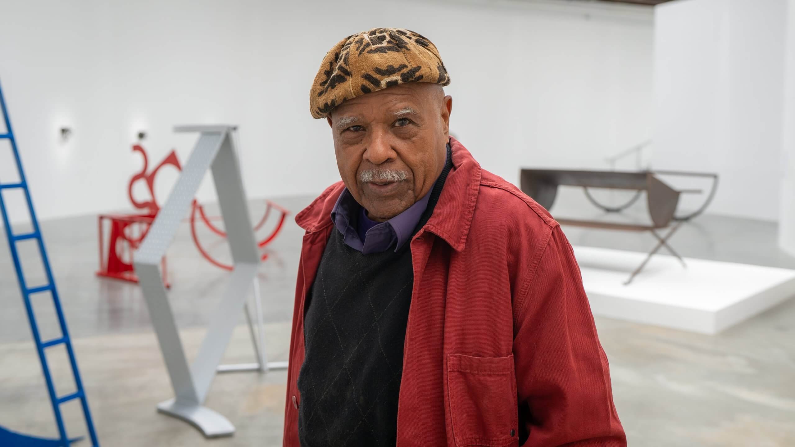 Un homme âgé portant un béret à motifs, une veste rouge et un pull noir se tient dans une galerie d'art avec des sculptures modernes en arrière-plan.