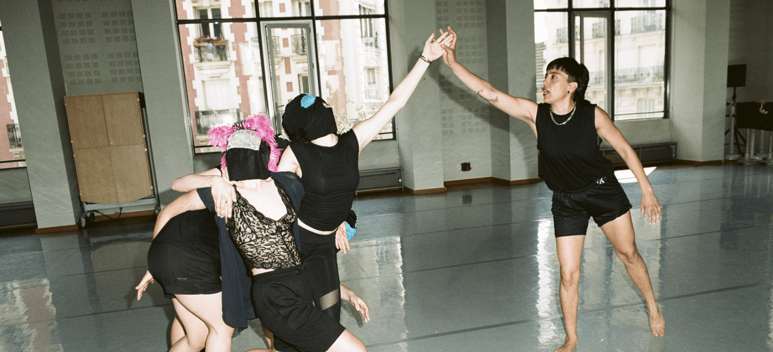 Quatre personnes portant des tenues noires et des couvre-chefs se regroupent tandis qu'une cinquième personne, le bras levé, se tient à l'écart dans un studio de danse spacieux, éclairé par le soleil et doté de grandes fenêtres.