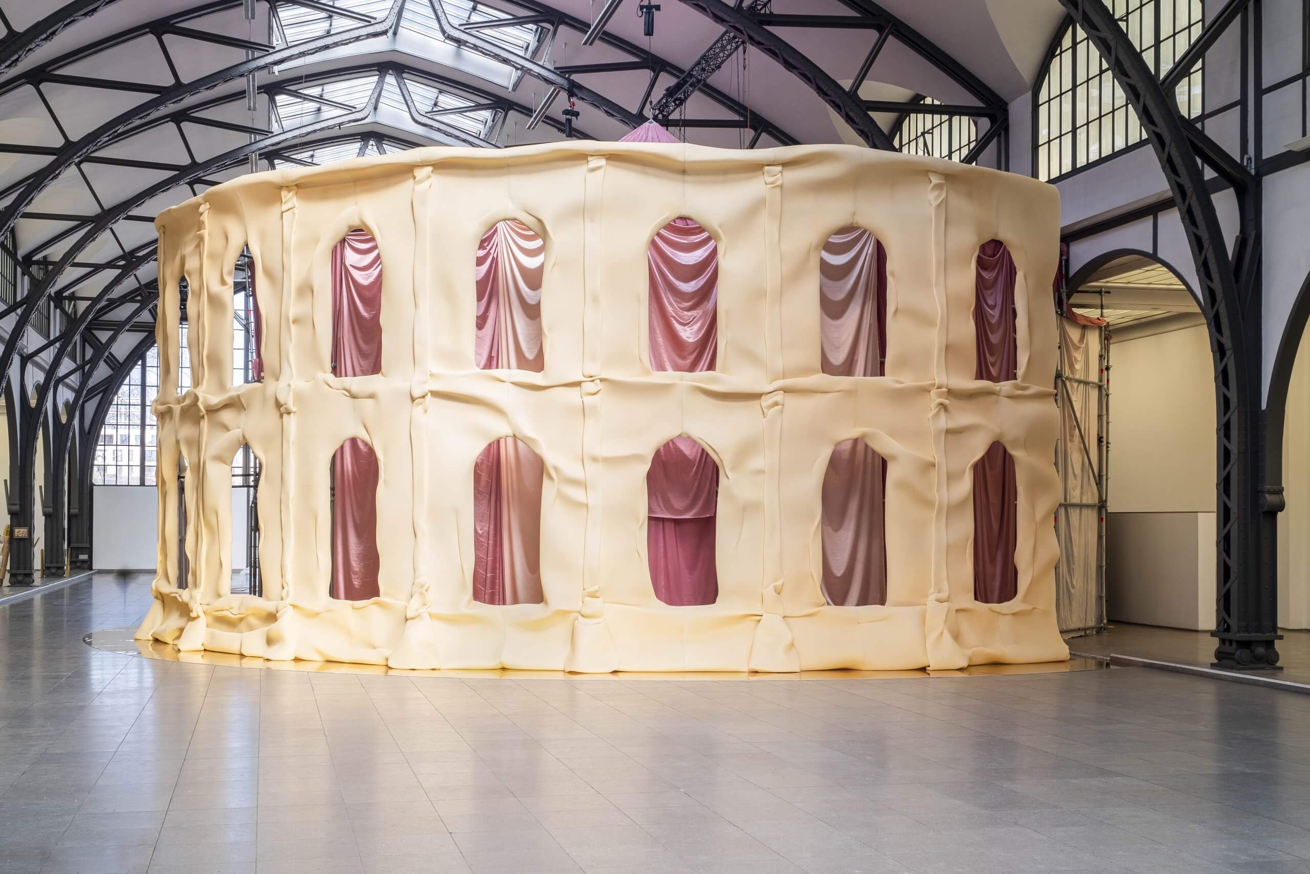 Une grande structure recouverte de tissu, ressemblant au Colisée romain, se trouve à l'intérieur d'une galerie de style industriel avec un haut plafond vitré.