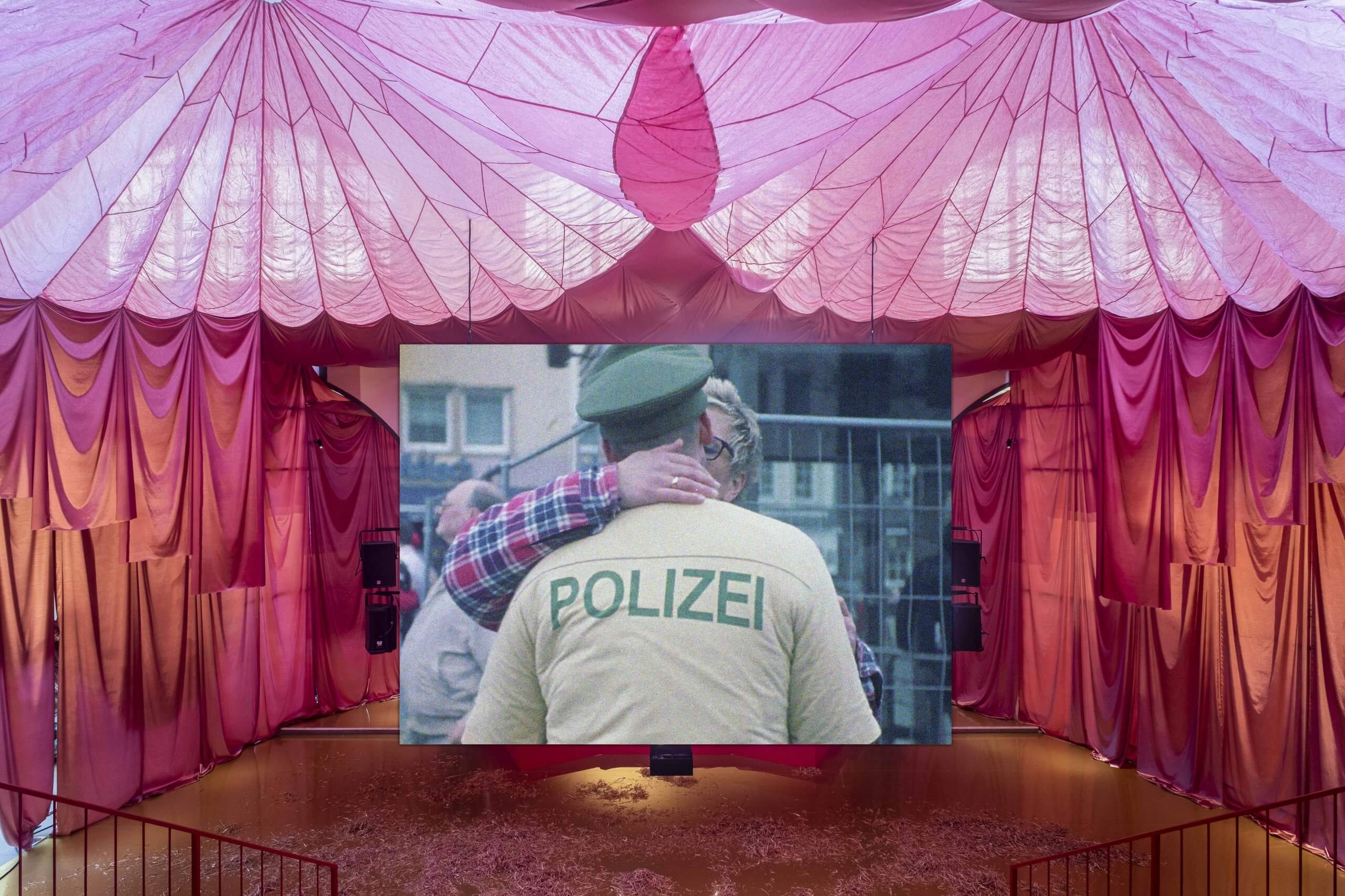 Une pièce drapée de tissu rose avec un écran central affichant une photo de deux personnes s'embrassant, l'une portant une veste étiquetée Polizei.