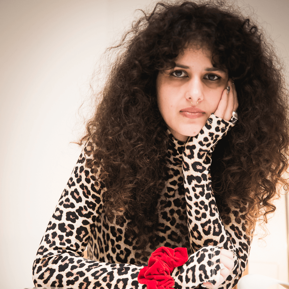 Une personne aux longs cheveux bouclés et aux yeux maquillés de couleur sombre porte un haut imprimé léopard et repose sa tête sur sa main avec un chouchou rouge au poignet.