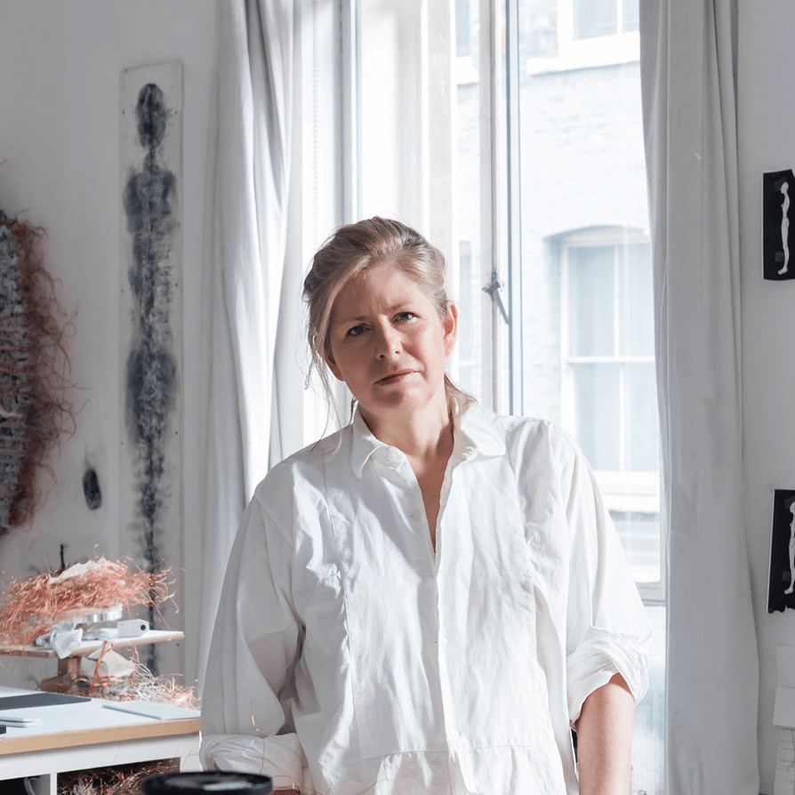 Une femme en chemise blanche se tient dans une pièce lumineuse avec des rideaux blancs, des œuvres d'art et divers matériaux visibles à l'arrière-plan.
