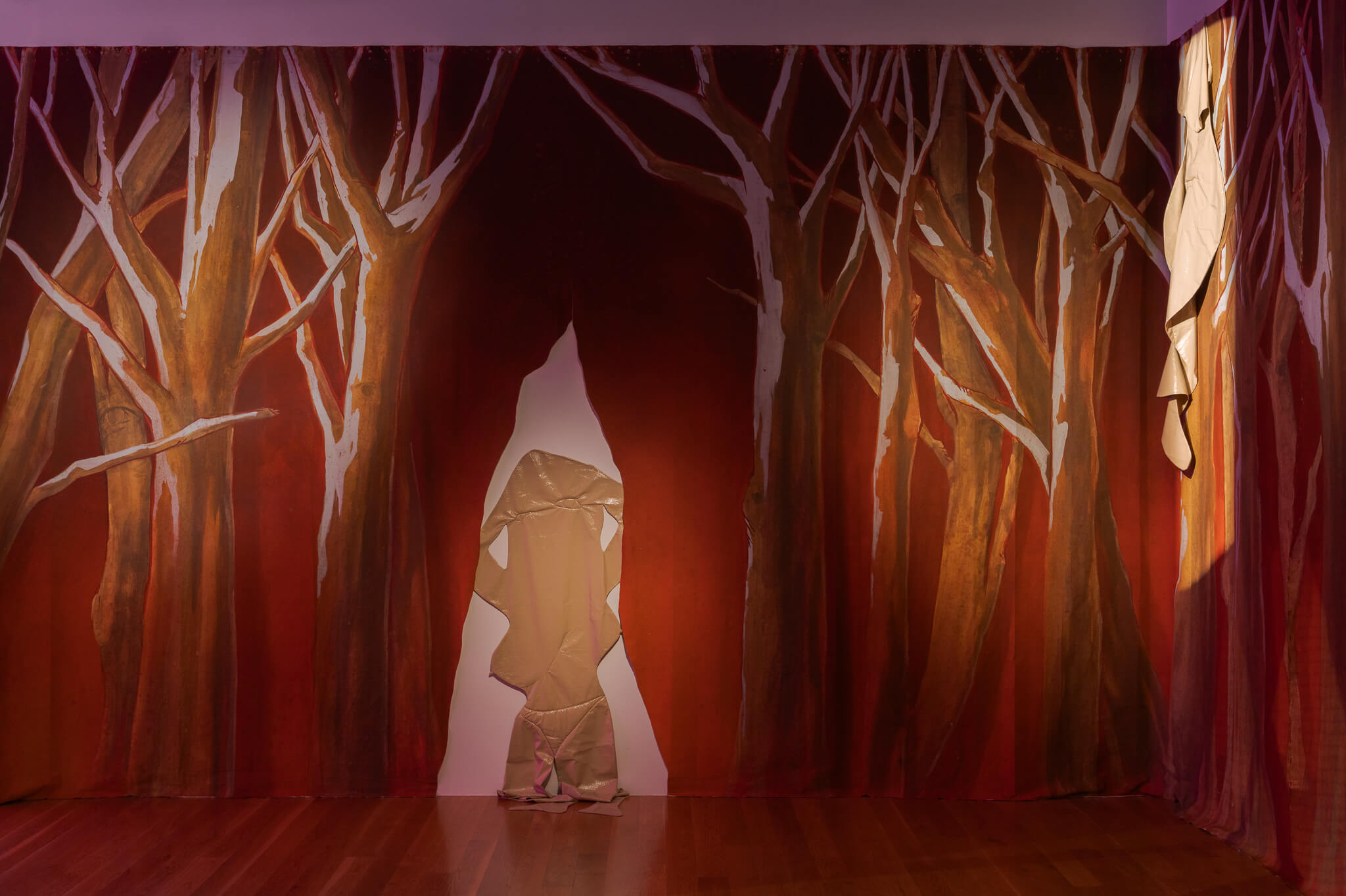 Des silhouettes d'arbres bruns sont peintes sur une draperie rouge ; un personnage en tissu beige se tient devant une fente dans le rideau, un autre morceau de tissu beige étant suspendu à droite.