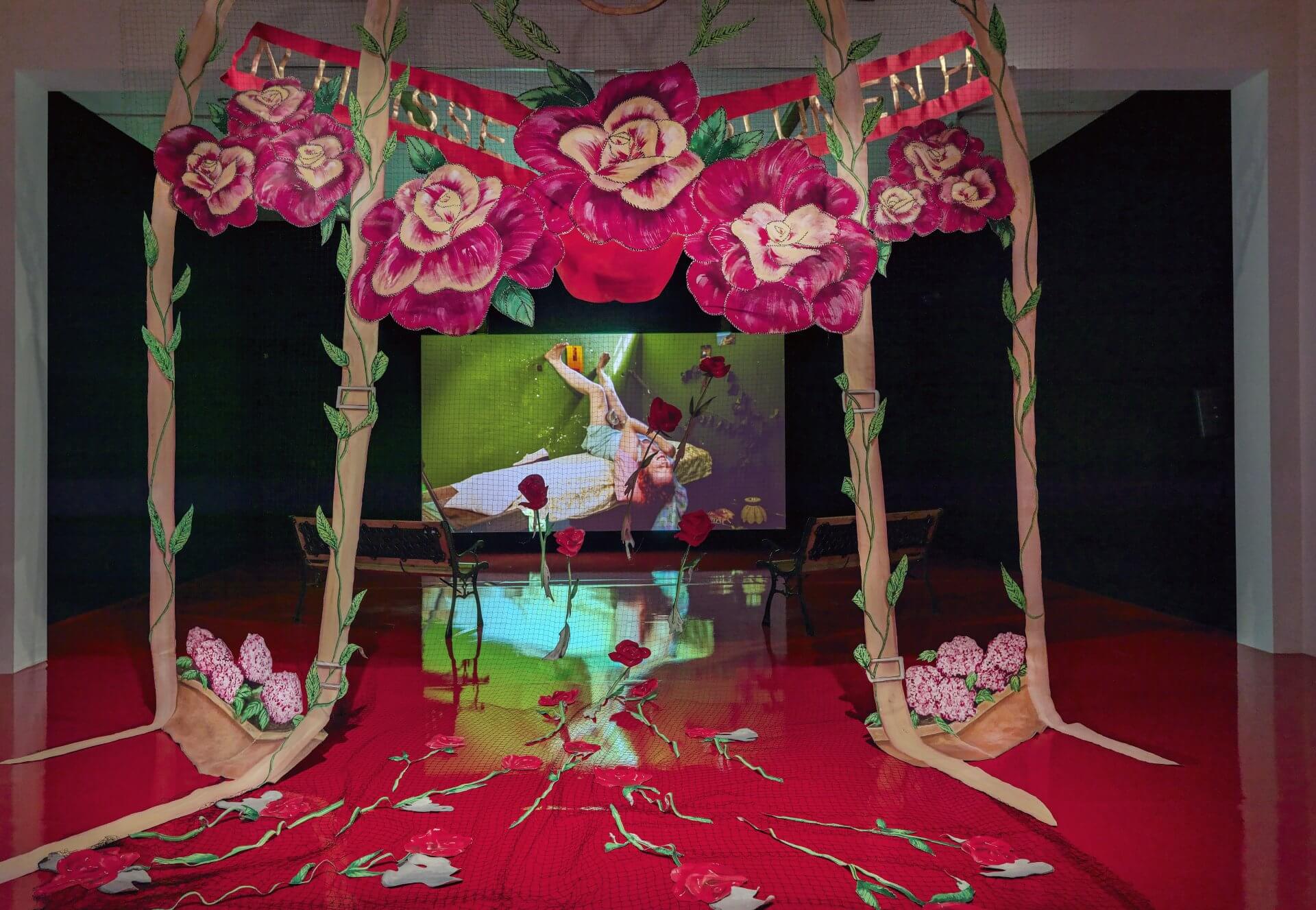 A decorated arch with large pink flowers frames a red floor scattered with petals; in the background, a screen displays a person in white reaching upward.