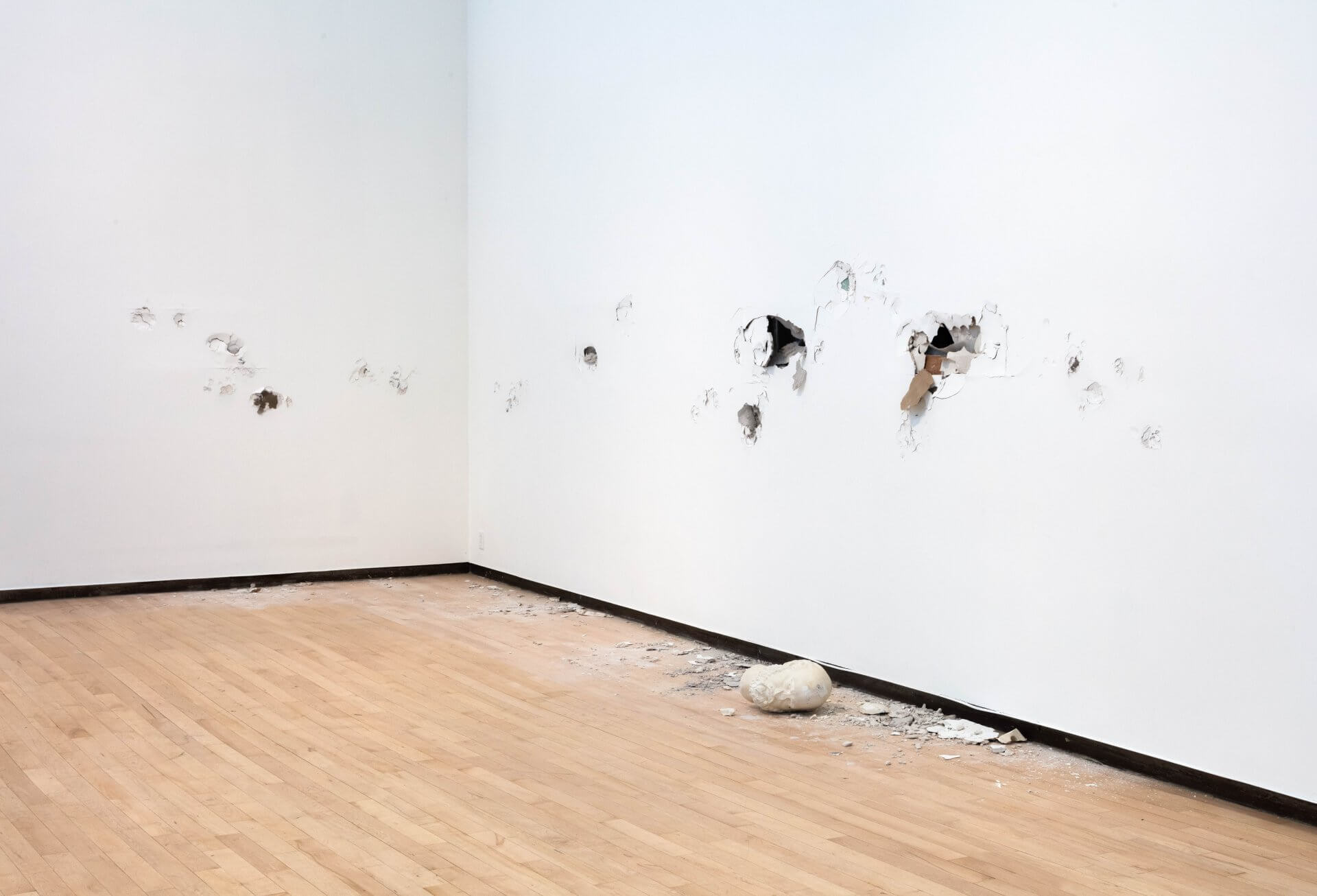 Un mur de galerie blanc avec plusieurs trous et dommages, des débris éparpillés sur le sol en bois, et un grand objet ressemblant à un rocher reposant près du mur.