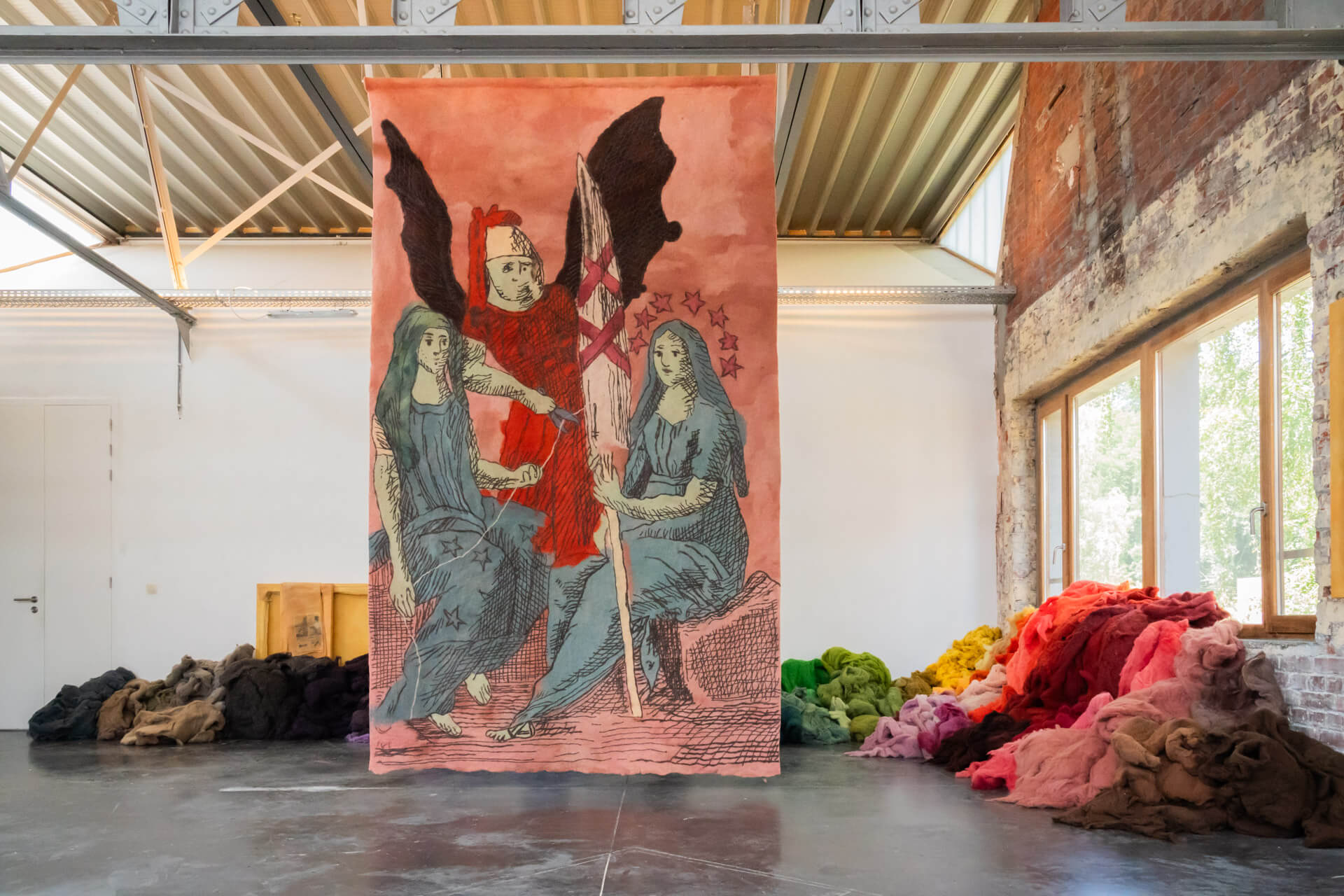 Une grande tapisserie illustrée représentant trois personnages de style médiéval est suspendue dans une galerie industrielle, avec des piles de tissus colorés sur le sol à proximité.